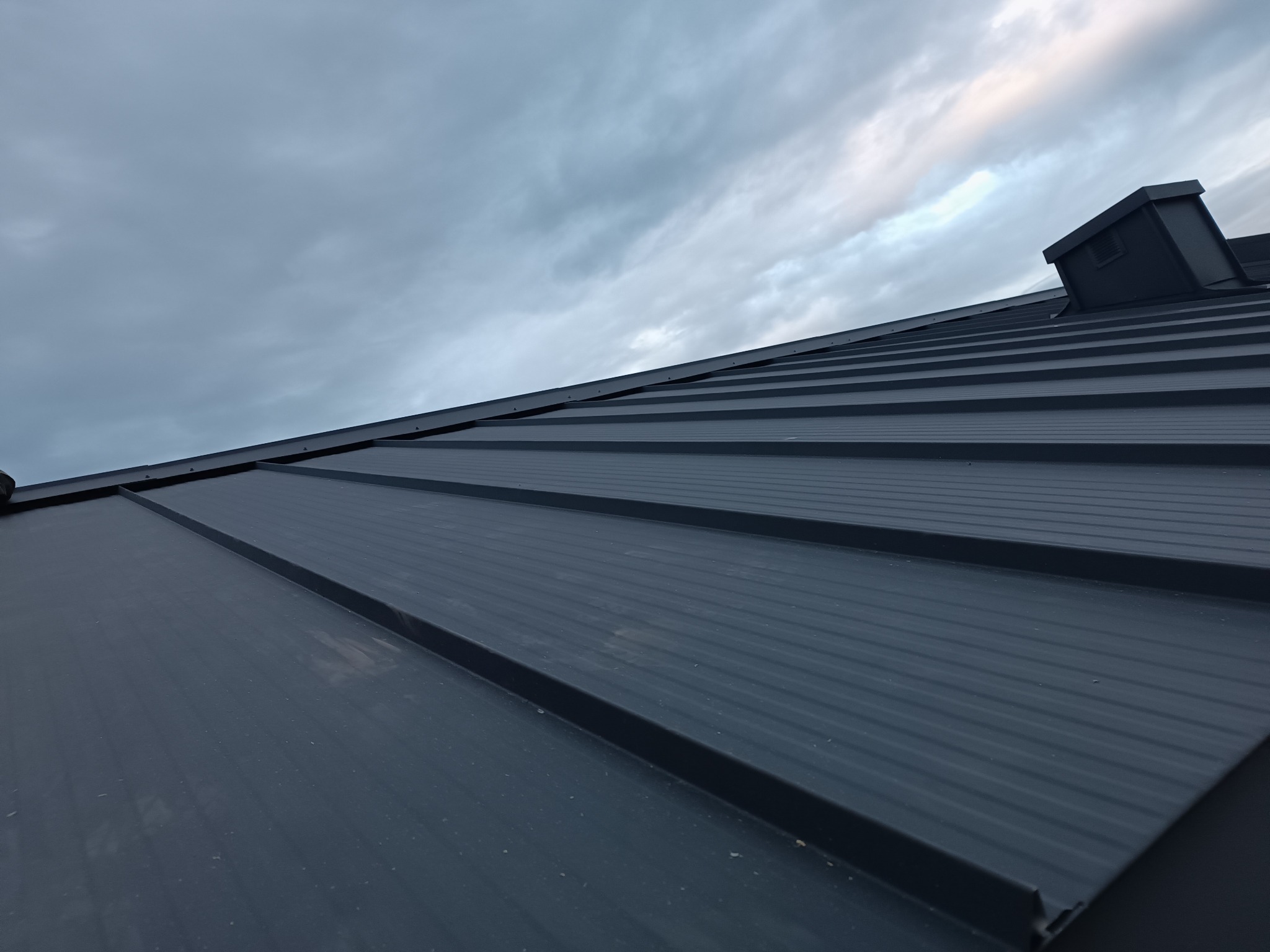 Nowoczesny, grafitowy dach z blachy na rąbek stojący, widziany pod kątem, z kominem w tle i zachmurzonym niebem. Minimalistyczny design.
