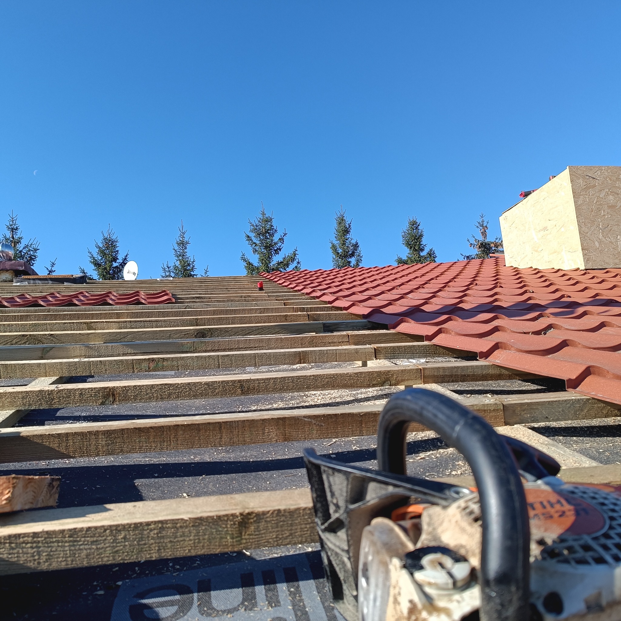 Częściowy widok na konstrukcję dachu z czerwoną dachówką, drewniane łaty i membranę, z piłą łańcuchową na pierwszym planie i błękitnym niebem w tle.