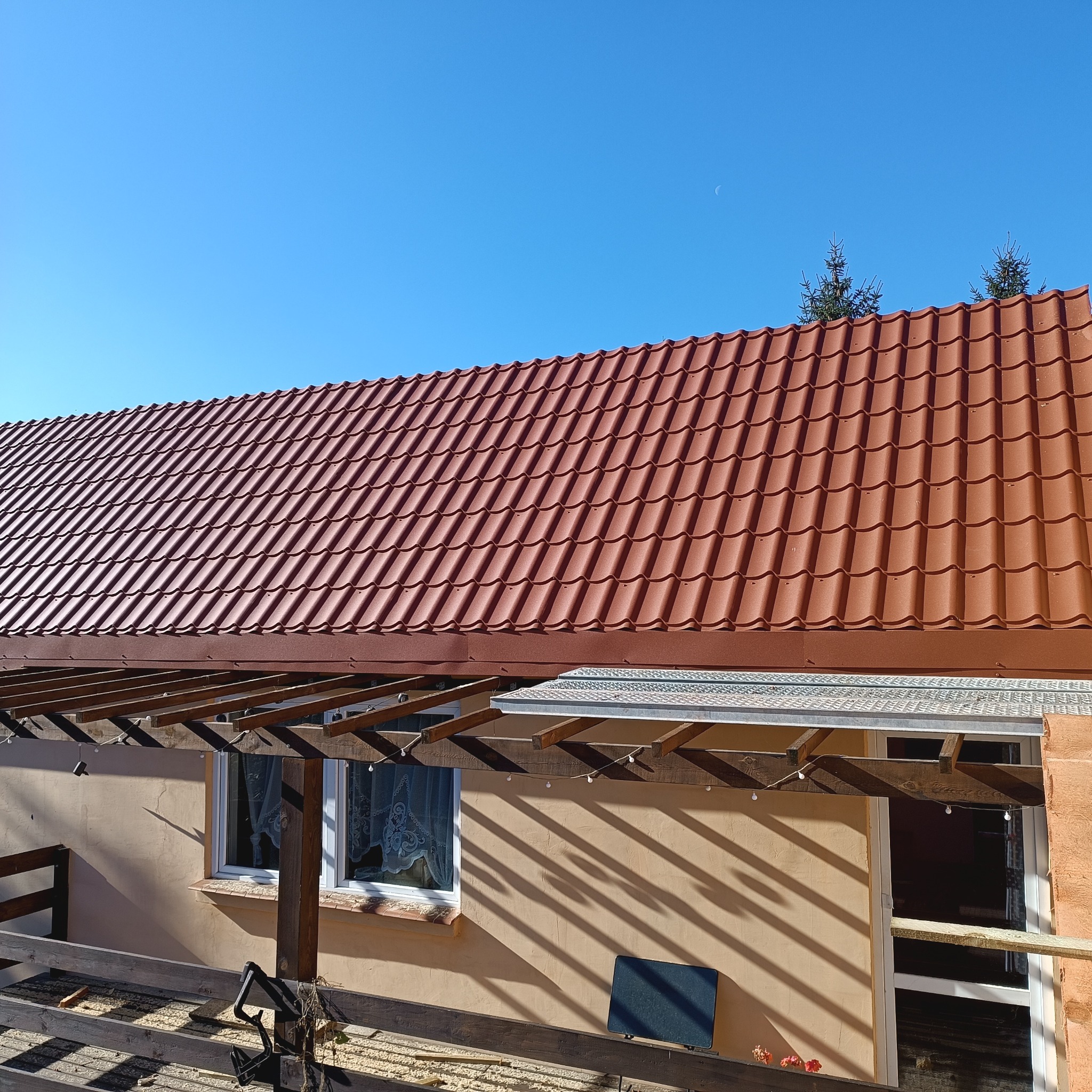 Nowy, brązowy dach z dachówki ceramicznej pod błękitnym niebem z widocznym sierpem księżyca. Ujęcie z dołu na zewnątrz budynku.