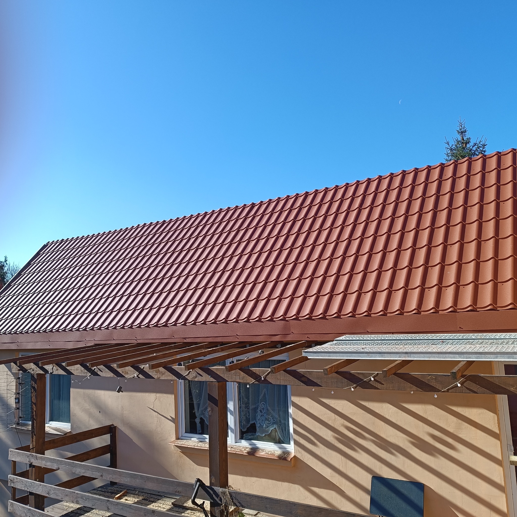 Czerwony dach z dachówki metalowej na budynku w słoneczny dzień. Widoczny fragment balkonu i błękitne niebo z małym księżycem w tle.