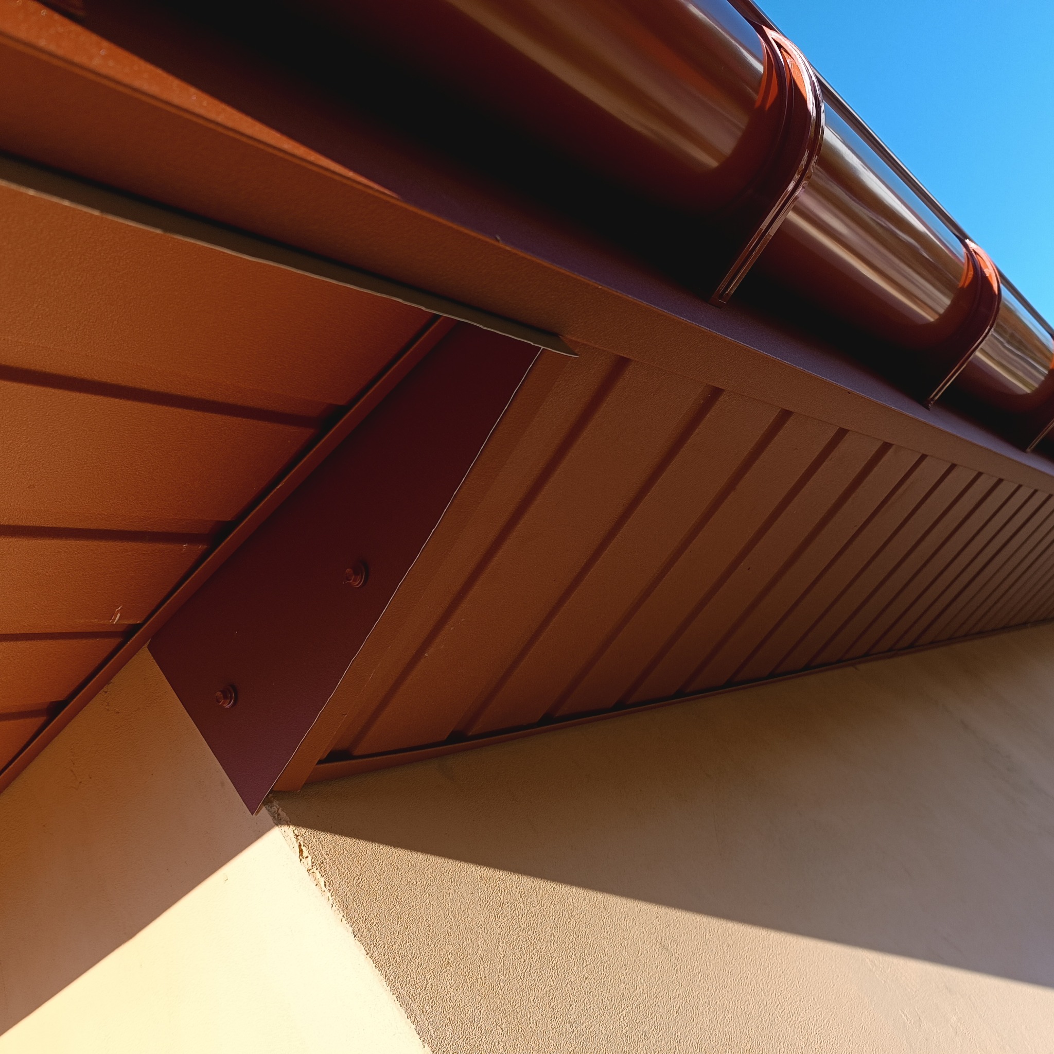 Brązowa rynna dachowa z podsufitką i fragmentem elewacji budynku w słoneczny dzień. Widok z dołu.