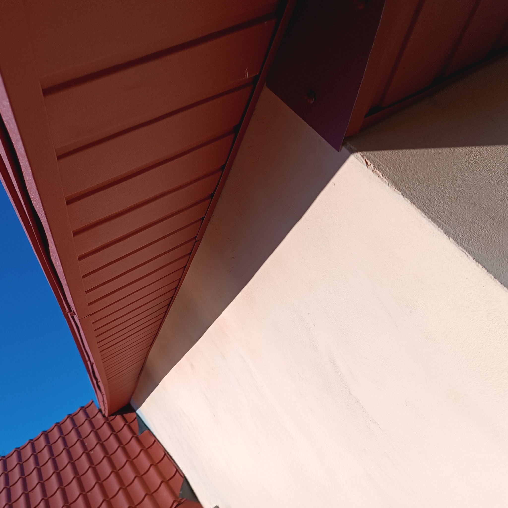 Fragment dachu z brązową podbitką i czerwoną dachówką, widziany z dołu pod kątem, na tle błękitnego nieba i beżowej ściany budynku.