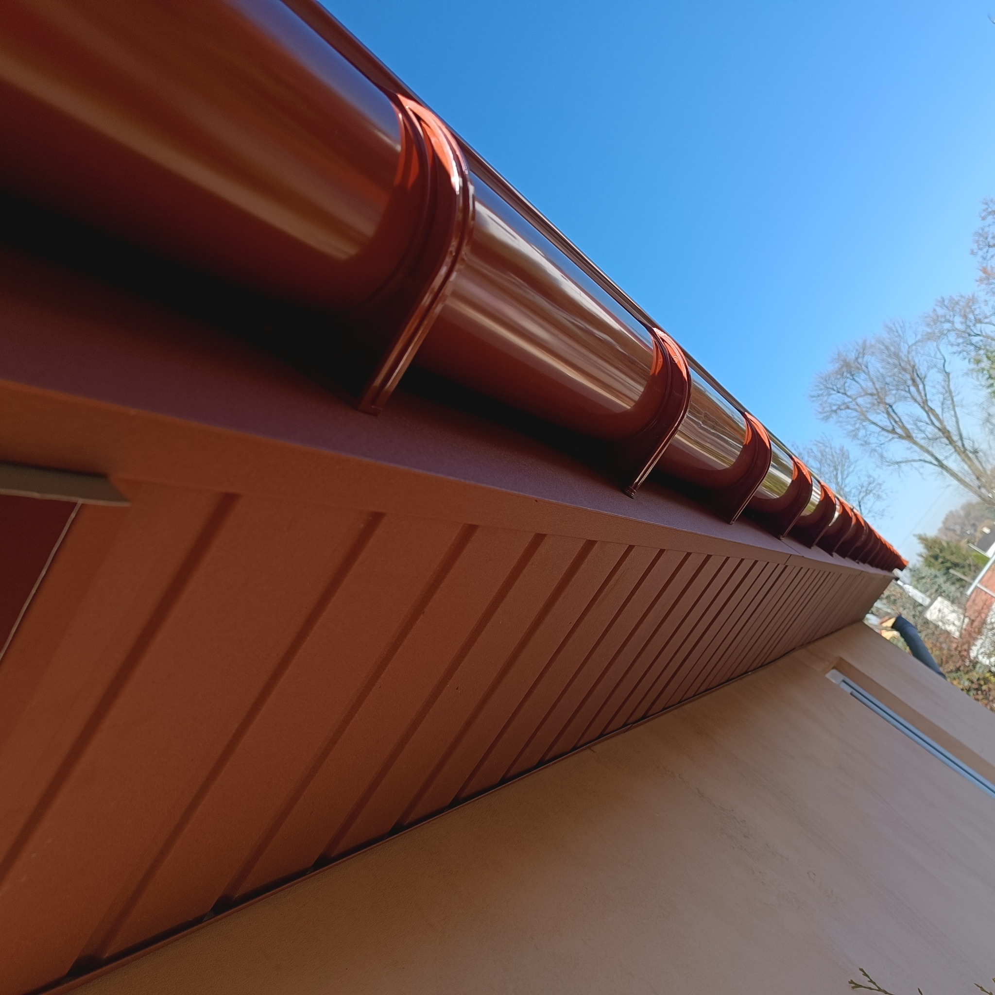 Brązowe rynny zamontowane na podbitce dachowej, widok z dołu na tle błękitnego nieba. Ujęcie podkreśla detale orynnowania i estetykę wykonania.