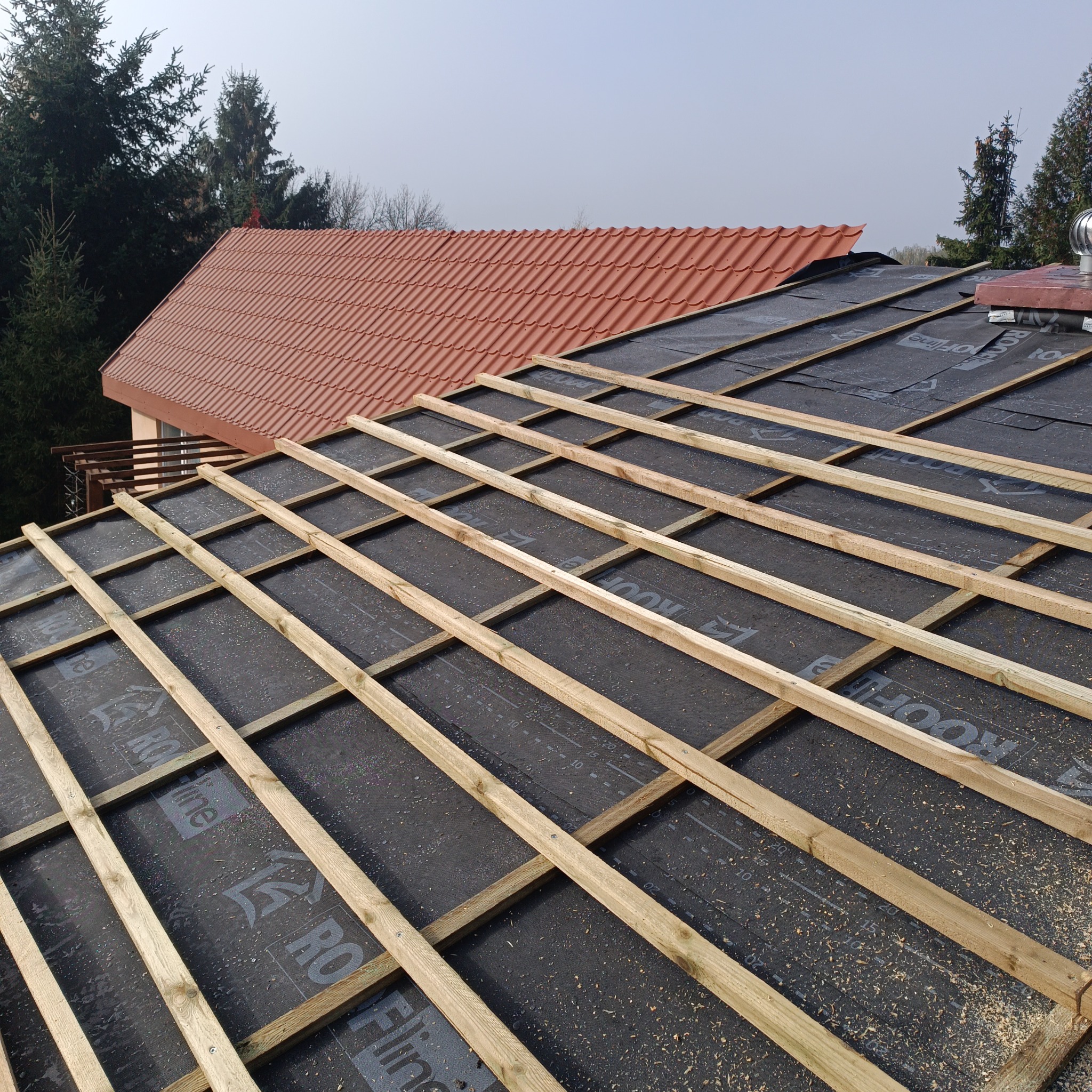 Więźba dachowa z łatami na czarnej membranie, fragment czerwonego dachu w tle. Widoczne drzewa i niebo. Etap budowy dachu z bliska.
