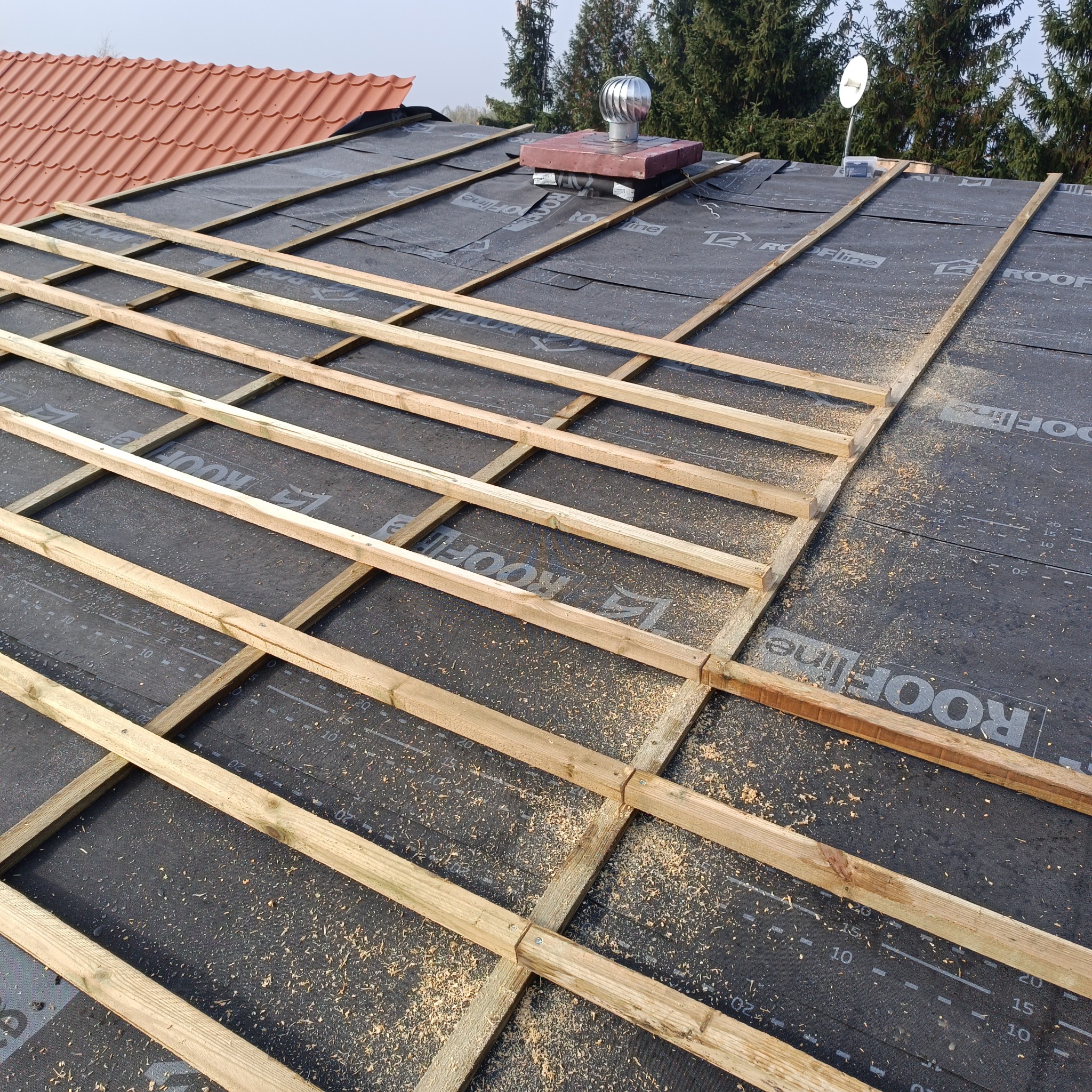 Montaż łat na membranie dachowej z widocznym kominkiem wentylacyjnym i anteną satelitarną w tle. Etap przygotowania pod krycie dachu. Wióry na powierzchni.