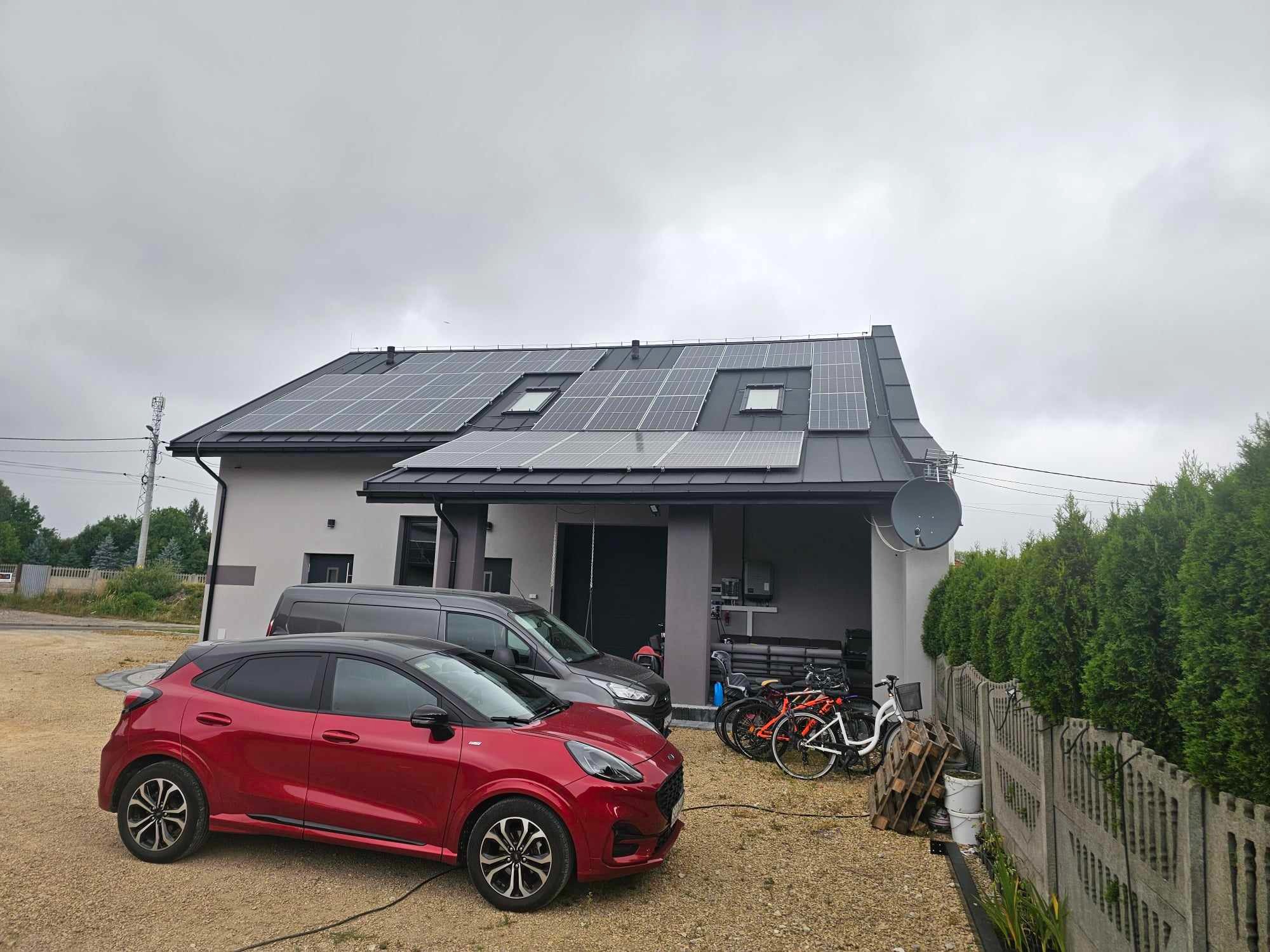 Szary dom jednorodzinny z panelami słonecznymi na dachu, widok z zewnątrz. Na podjeździe czerwony samochód i szary van.