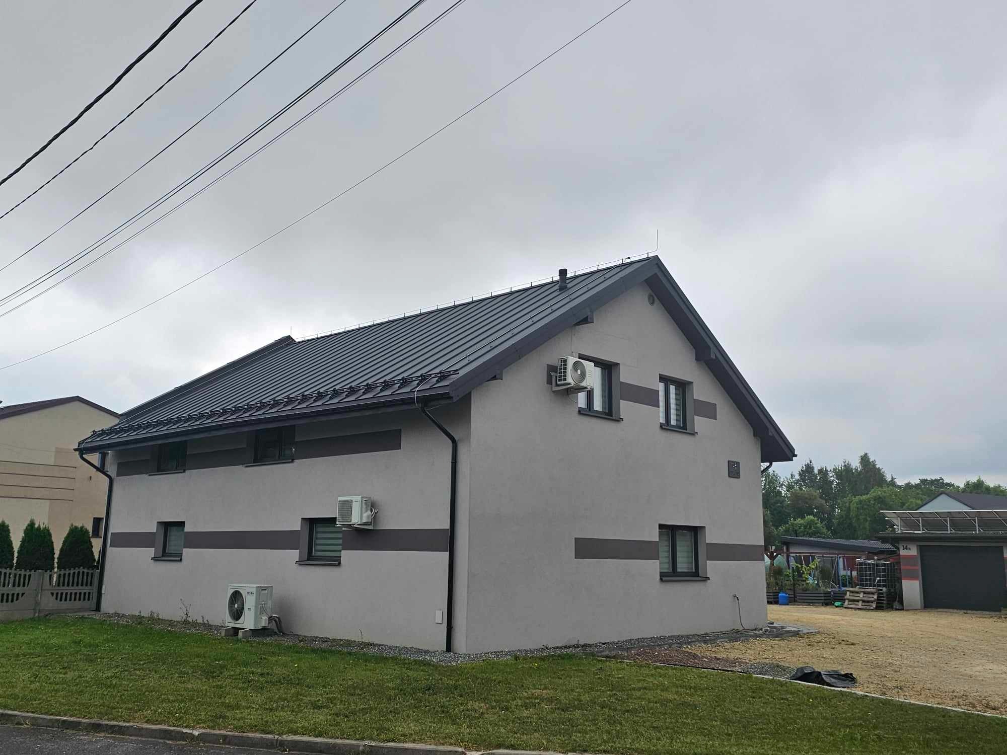 Szary dom jednorodzinny z ciemnoszarym dachem z blachy na rąbek stojący, z zamontowanymi klimatyzatorami na elewacji i rynnami w kolorze dachu.