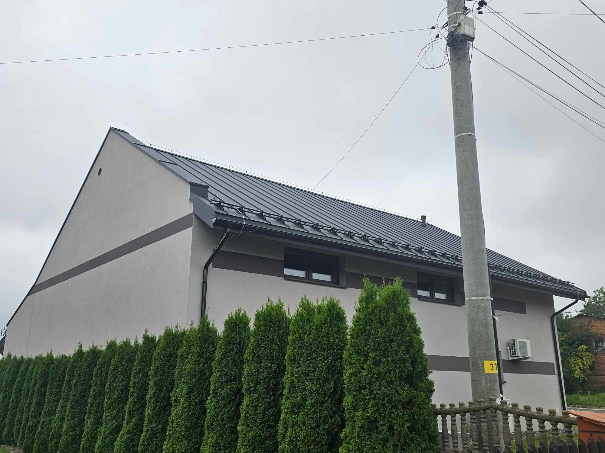 Szary dom jednorodzinny z ciemnym, metalowym dachem i rynnami. Wzdłuż ogrodzenia rosną wysokie tuje. Słup energetyczny z przewodami w tle.