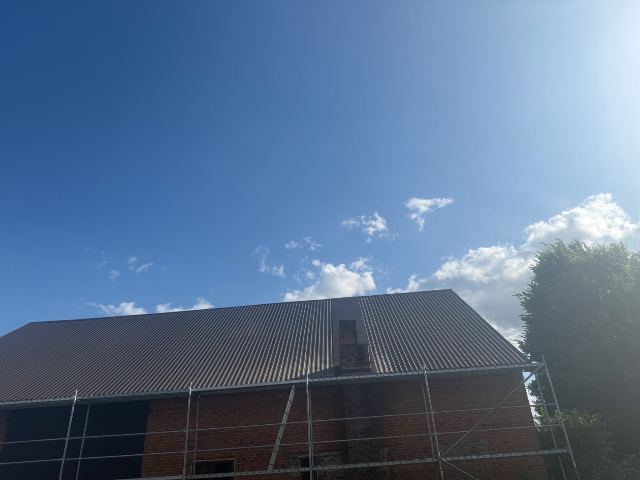 Ceglany budynek z nowym, brązowym dachem z blachy trapezowej w trakcie montażu, widoczne rusztowanie. Częściowo błękitne niebo z chmurami w tle.