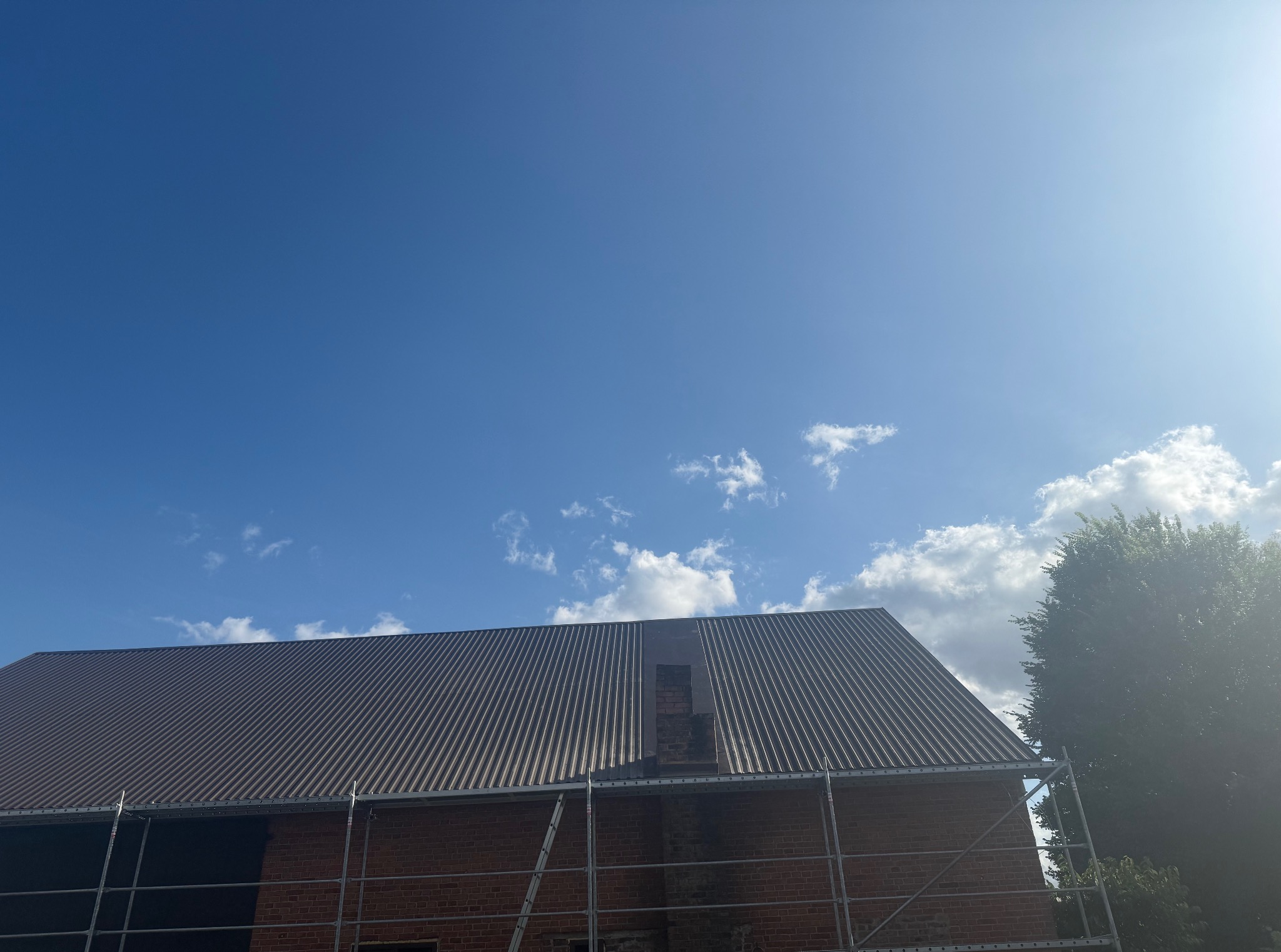 Częściowo pokryty blachą falistą dach z kominem na tle błękitnego nieba z chmurami. Widoczne rusztowanie i fragment drzewa po prawej stronie.