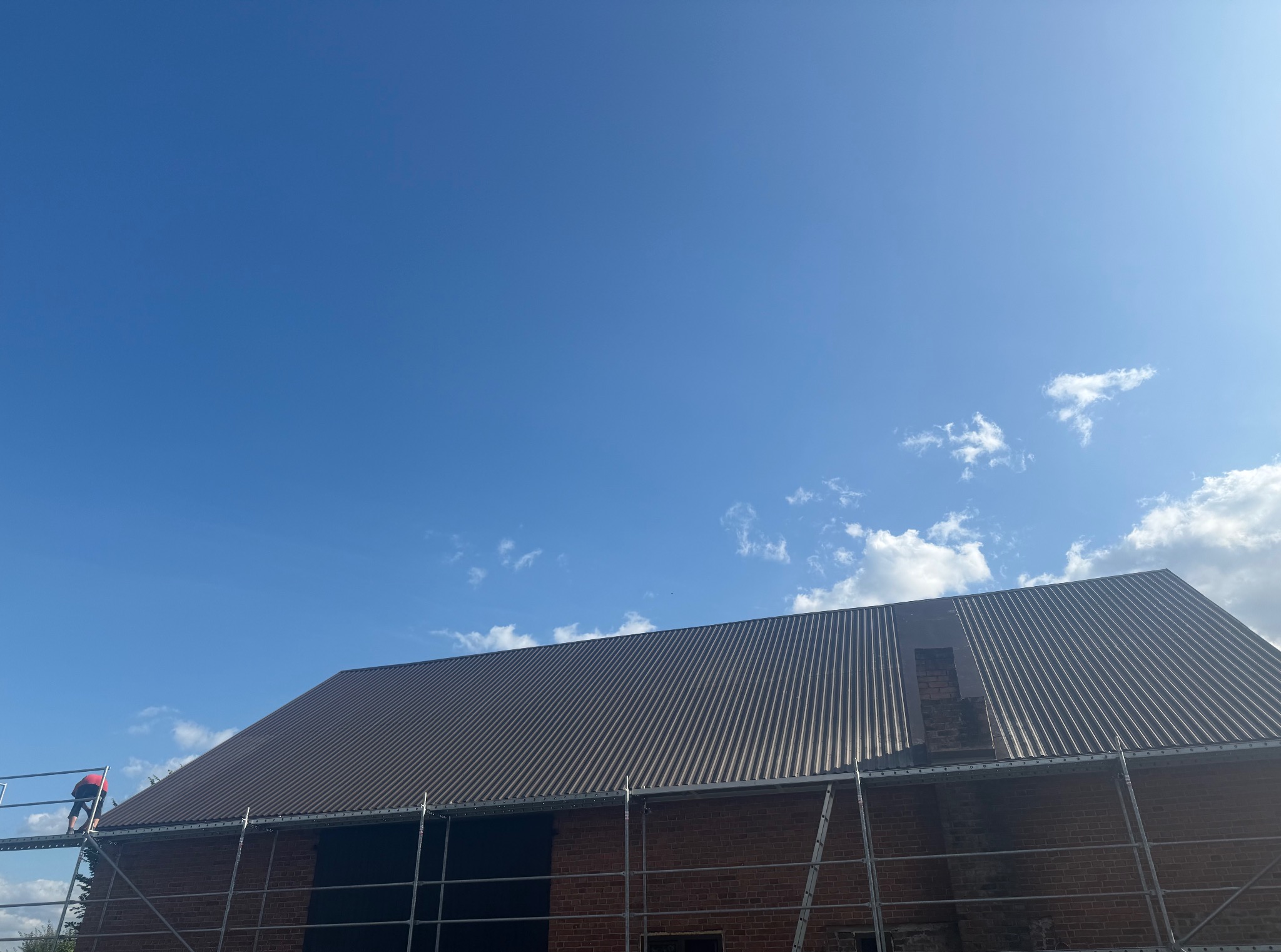 Ciemnobrązowy dach z blachy falistej na ceglanym budynku, widziany z rusztowania na tle błękitnego nieba z chmurami. Fragment komina w trakcie prac.