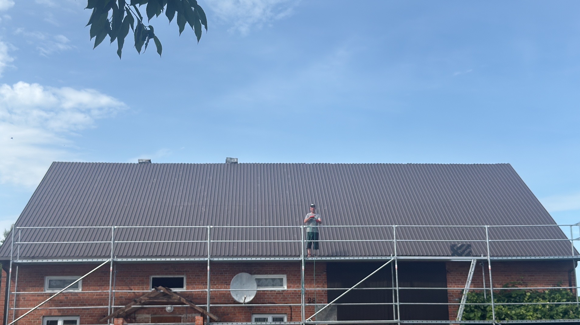 Nowy, brązowy dach z blachy falistej na ceglanym budynku. Na dachu stoi dekarz na rusztowaniu. Widoczne liście drzewa i błękitne niebo.
