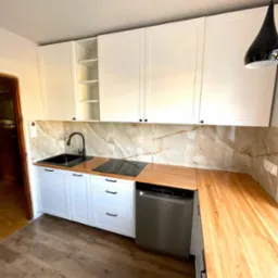 Nowoczesna kuchnia z białymi szafkami, drewnianym blatem i czarną armaturą, marmurowe płytki na ścianie i drewniana podłoga.