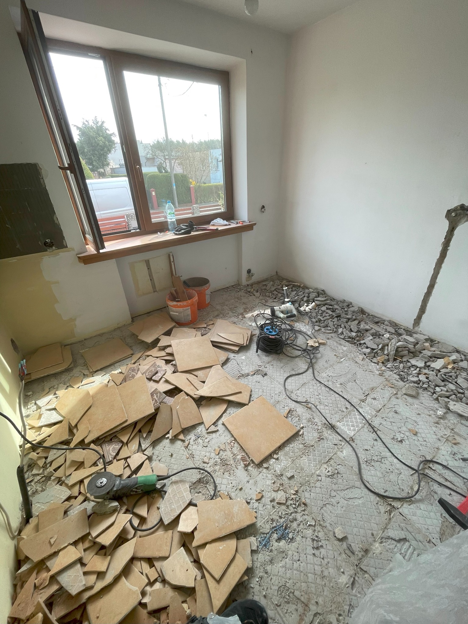 Pokój w trakcie skuwania starych płytek podłogowych, widoczne resztki gruzu, narzędzia i fragmenty płytek, okno z widokiem na zewnątrz.