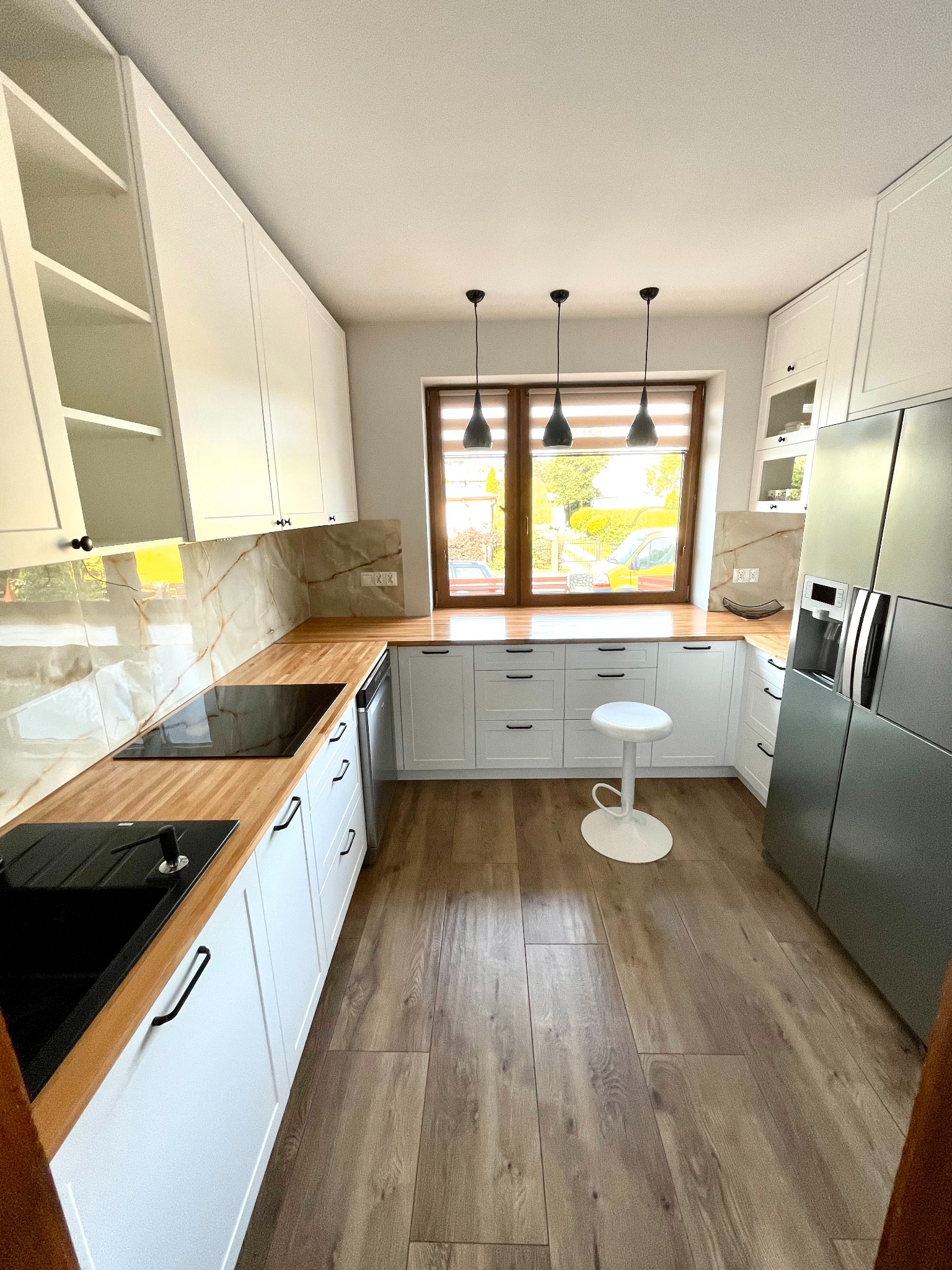 Jasna kuchnia z drewnianymi blatami, białymi szafkami z czarnymi uchwytami, marmurowym backsplashiem i drewnianą podłogą. Nad oknem wiszą trzy czarne lampy. Biały stołek barowy stoi na środku...