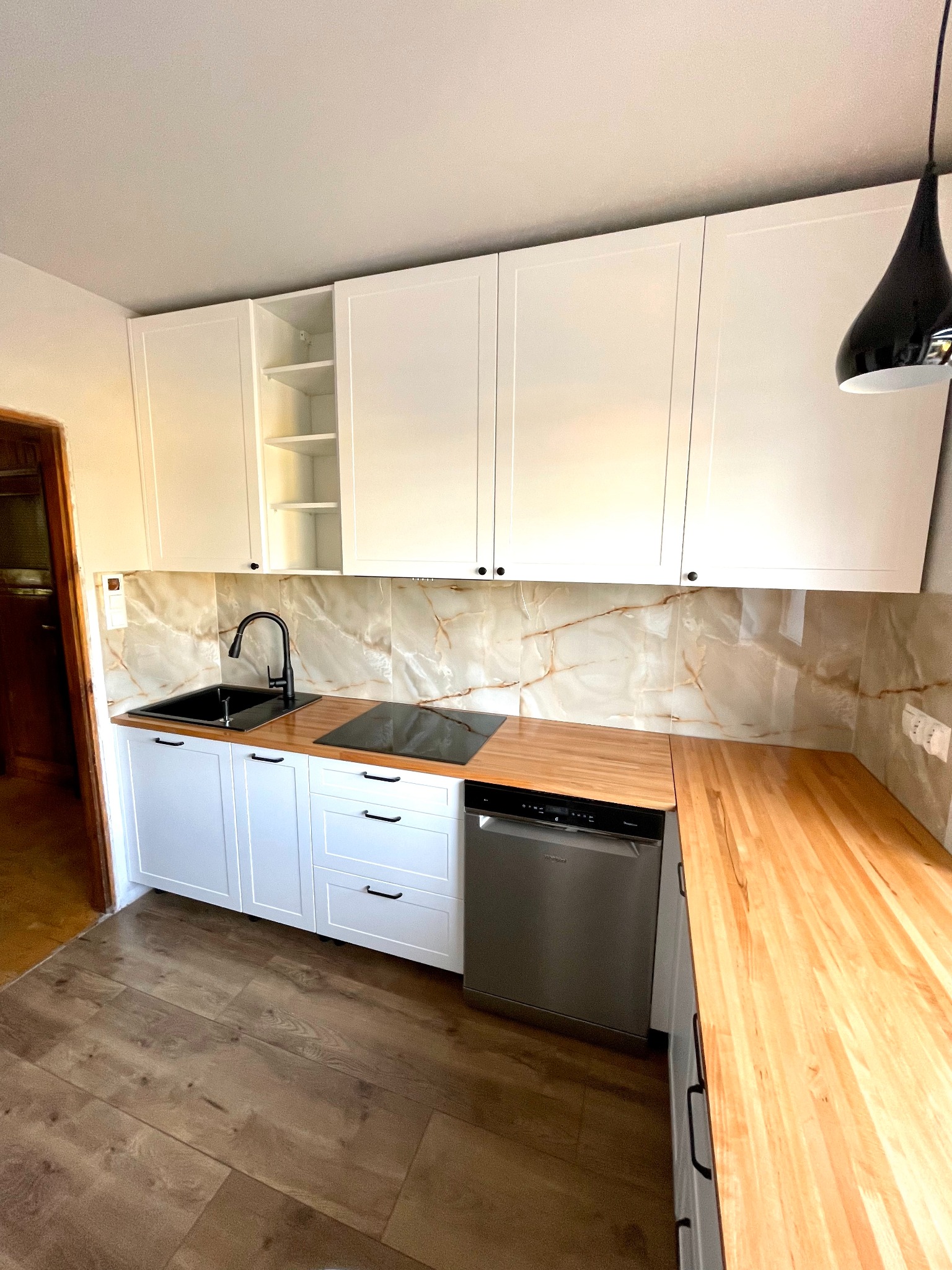 Nowoczesna kuchnia z białymi szafkami, drewnianym blatem i czarną armaturą, marmurowe płytki na ścianie i drewniana podłoga.