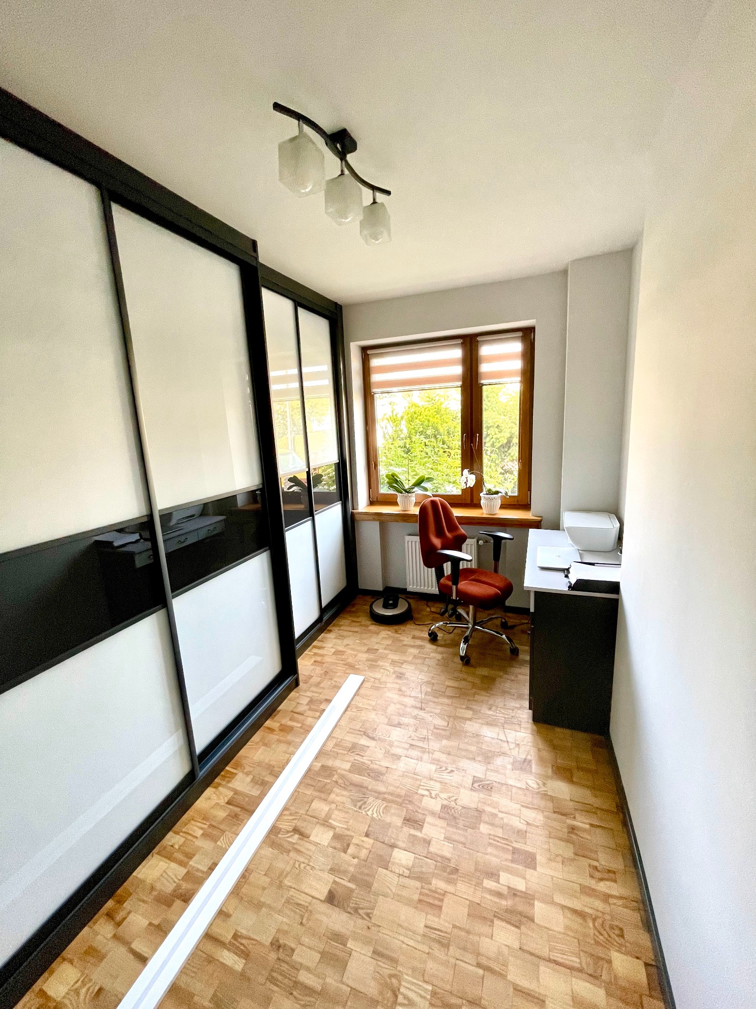 Pokój z dużym przesuwnym szafą z czarno-białymi frontami, biurkiem przy oknie, krzesłem biurowym i parkietem w jodełkę; widoczna listwa przypodłogowa przed szafą.