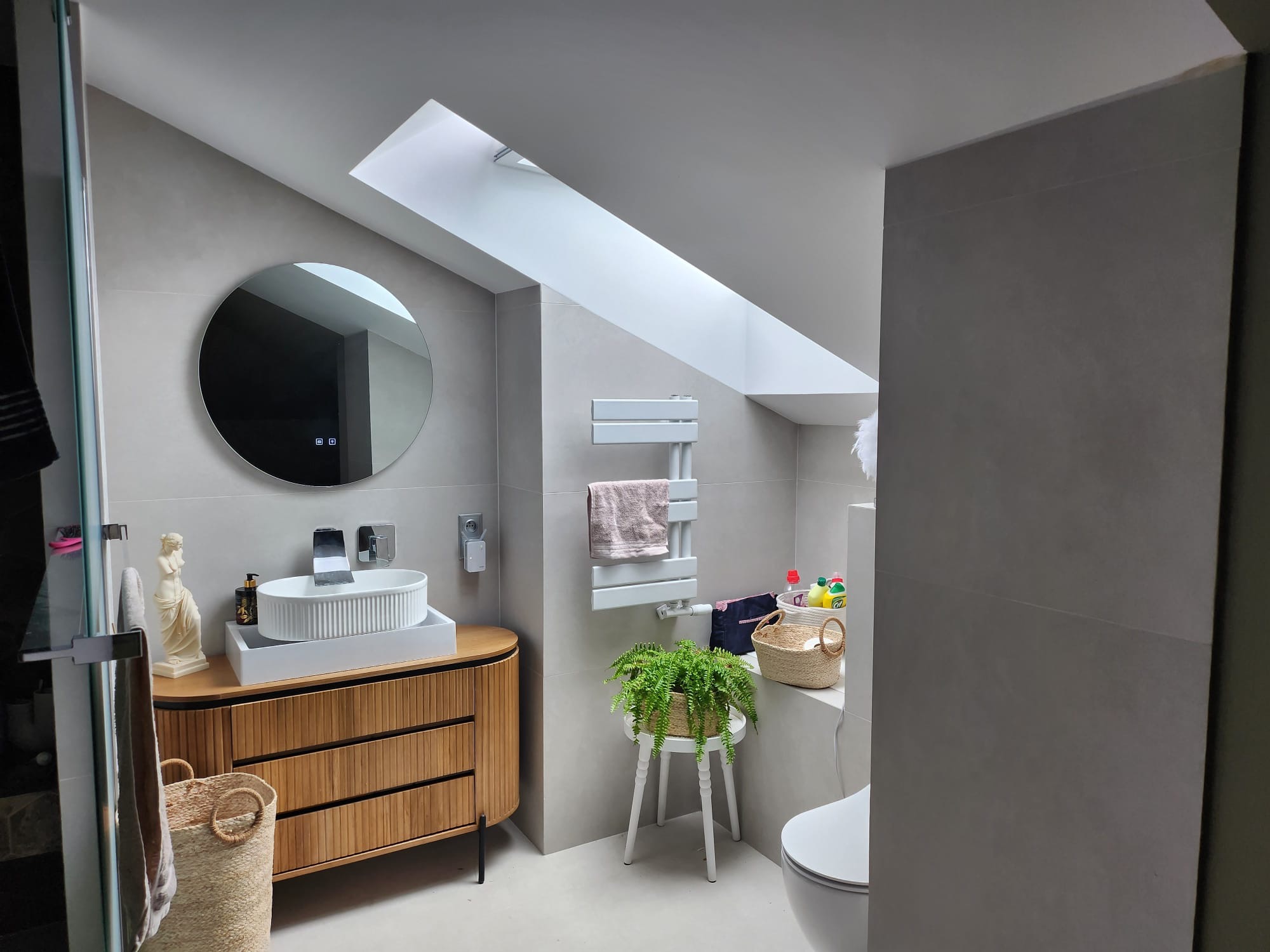 Nowoczesna łazienka na poddaszu z owalną umywalką nablatową, drewnianą szafką z lamelami, okrągłym lustrem z podświetleniem LED i grzejnikiem drabinkowym, doświetlona oknem dachowym.