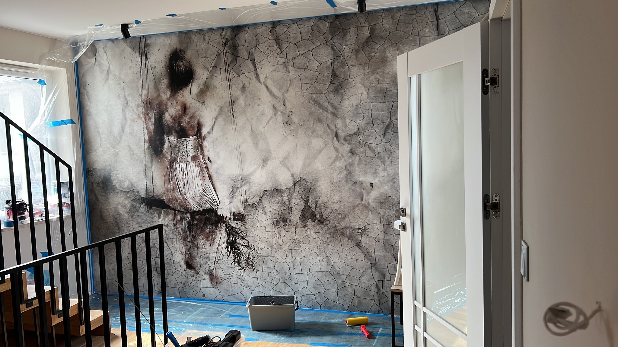 Ściana z fototapetą przedstawiającą postać kobiety na tle spękanej powierzchni, zabezpieczona folią malarską, widoczne narzędzia malarskie i fragment schodów.