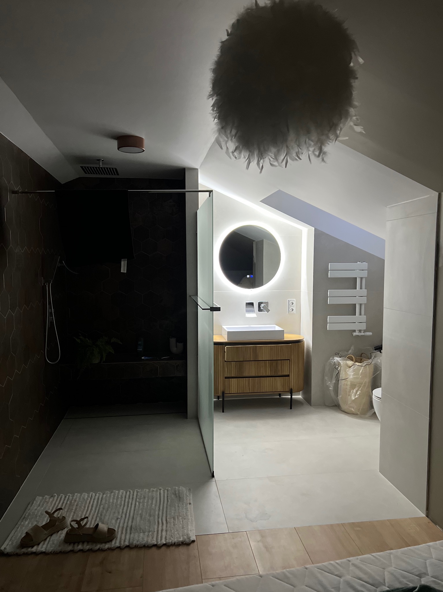 Nowoczesna łazienka na poddaszu z kabiną prysznicową wyłożoną heksagonalnymi płytkami, okrągłym lustrem z podświetleniem LED, drewnianą szafką pod umywalkę i grzejnikiem drabinkowym. Na podłodze...