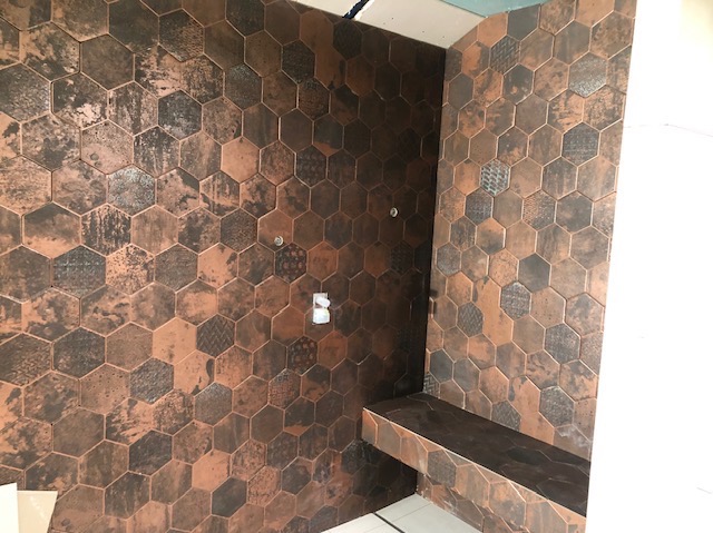 Kabina prysznicowa wyłożona heksagonalnymi płytkami w odcieniach miedzi i brązu z efektem postarzenia, widoczny fragment siedziska z tego samego materiału, dwa punkty mocowania armatury na ścianie.