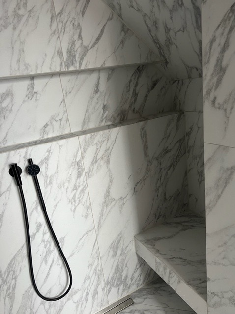 Nowoczesna kabina prysznicowa wykończona białym marmurem z szarymi żyłami, z wbudowanymi półkami i siedziskiem, widoczny czarny wąż natryskowy z dyszą.