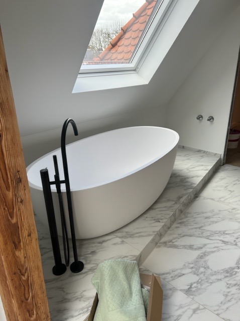 Nowoczesna, wolnostojąca wanna owalna z czarną armaturą w łazience na poddaszu z marmurową posadzką i oknem dachowym.