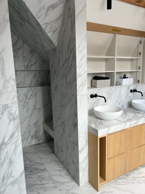 Nowoczesna łazienka z kabiną prysznicową wyłożoną płytkami imitującymi marmur, umywalki nablatowe na szafce z drewnianymi lamelami, białe półki w zabudowie.