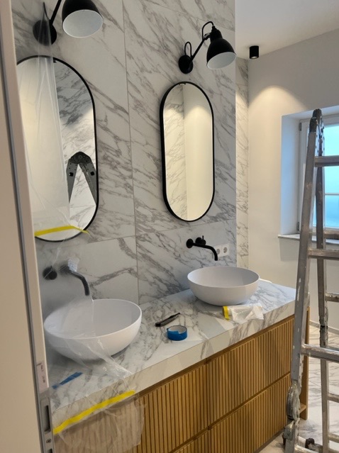 Dwie owalne umywalki nablatowe w nowoczesnej łazience z marmurowymi płytkami, czarną armaturą i oświetleniem, zabezpieczone folią malarską podczas prac wykończeniowych, z widoczną drabiną w tle.