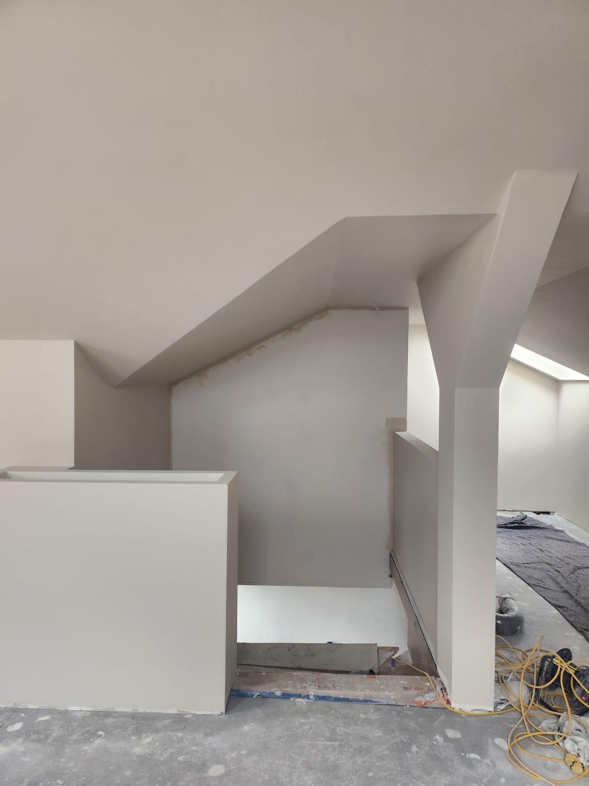 Wnętrze poddasza w trakcie prac wykończeniowych, widoczne świeżo pomalowane ściany i sufit, zabezpieczona podłoga, widoczne przewody elektryczne i schody w stanie surowym.