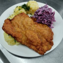 RANDEVU RIMMA HOLUBCHYKOVA, ANDRIY SOKALO SPÓŁKA CYWILNA - Złocisty sznycel na białym talerzu z ziemniakami i surówką z czerwonej kapusty, udekorowany koperkiem. Zdjęcie zrobione z bliska, widoczna struktura panierki.