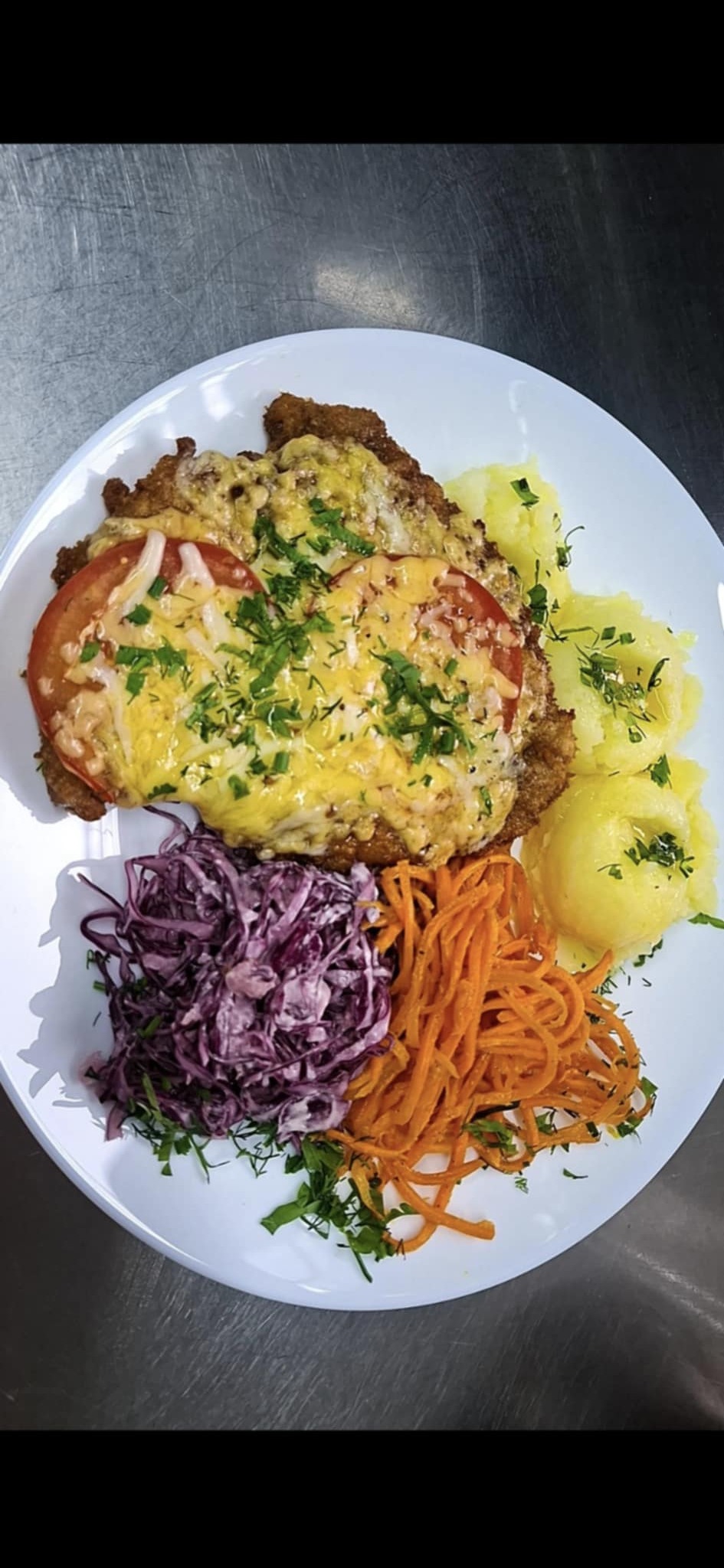 Sznycel zapiekany z serem i pomidorem na białym talerzu, z dodatkiem puree ziemniaczanego, surówki z czerwonej kapusty i marchewki, posypany świeżymi ziołami.
