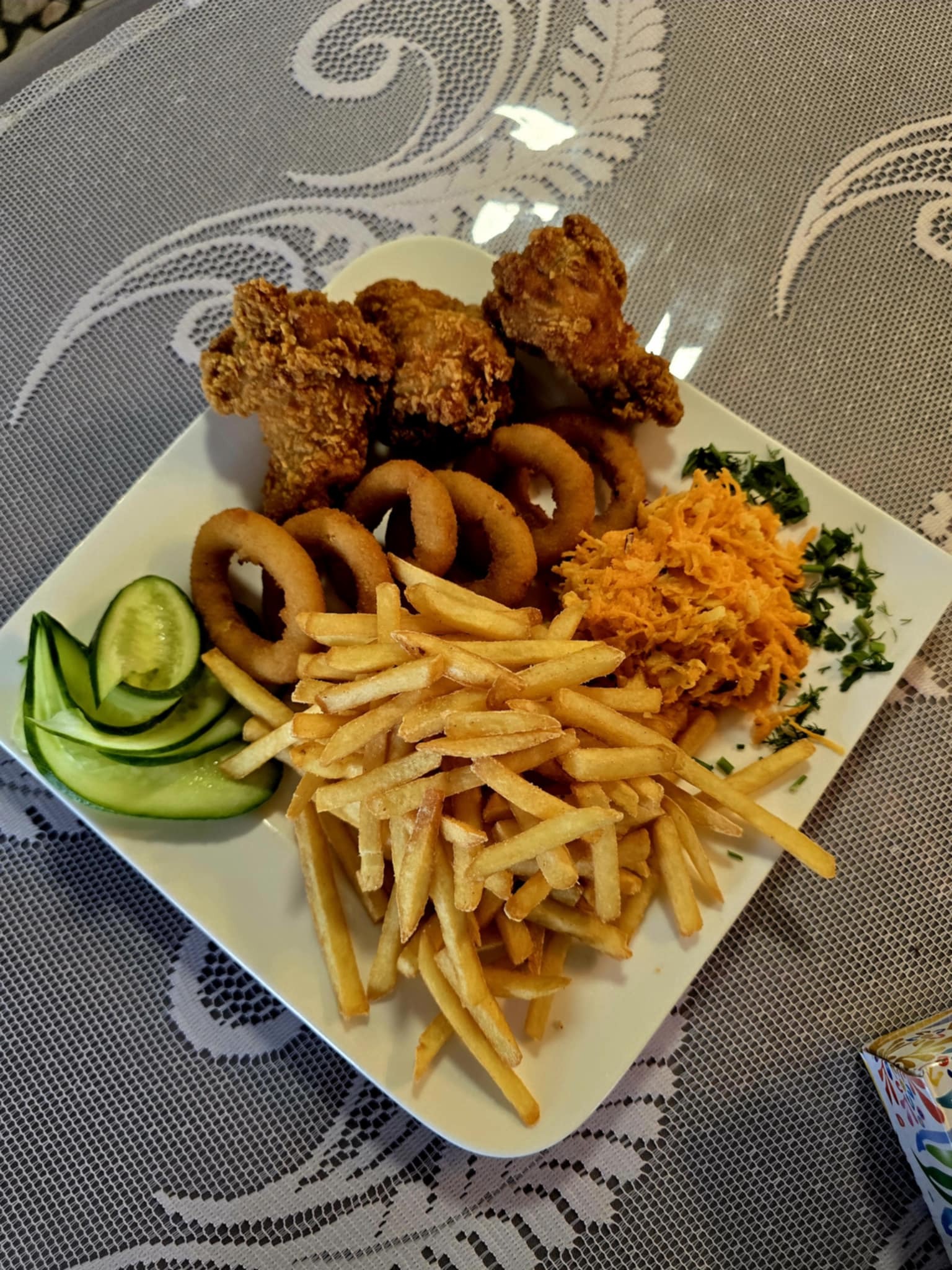 Kwadratowy talerz z frytkami, krążkami cebulowymi, smażonym kurczakiem, marchewką i ogórkiem na wzorzystym obrusie. Danie gotowe do spożycia.