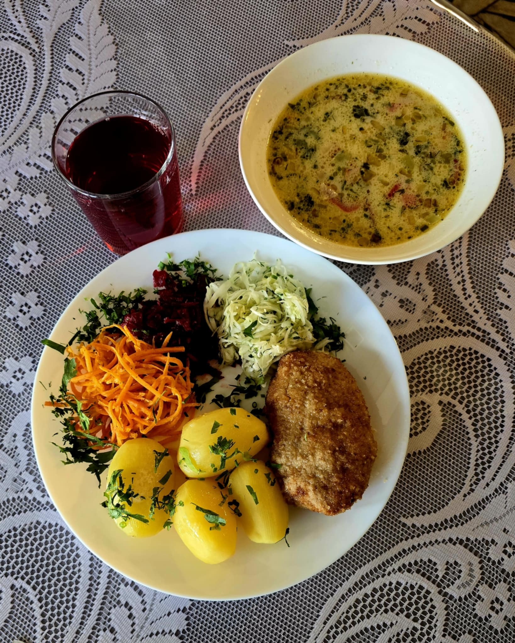 Kompletny obiad na białym talerzu: kotlet, ziemniaki z koperkiem, surówki, zupa w miseczce, szklanka soku na serwecie z koronkowym wzorem.