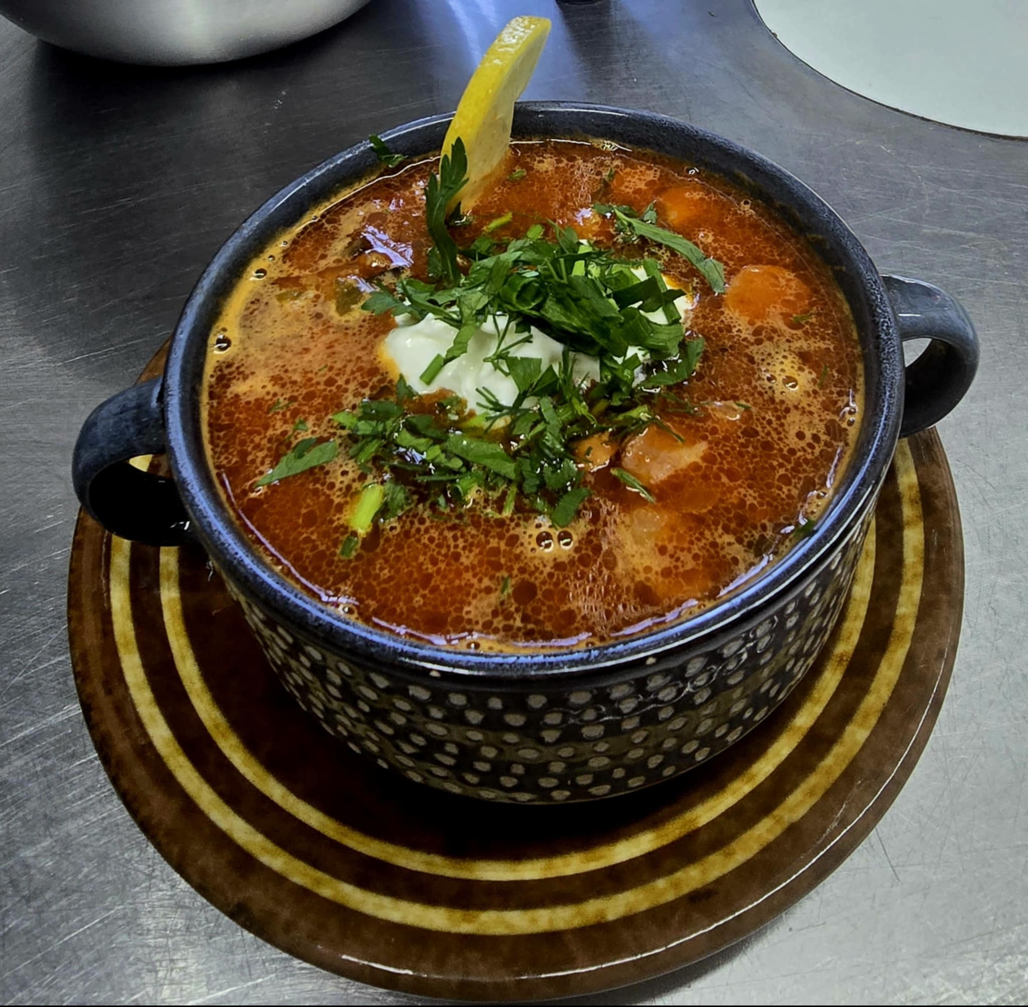 Aromatyczna zupa w czarnej misce, udekorowana kleksem śmietany, zieloną pietruszką i plasterkiem cytryny, na brązowym talerzu z paskami.