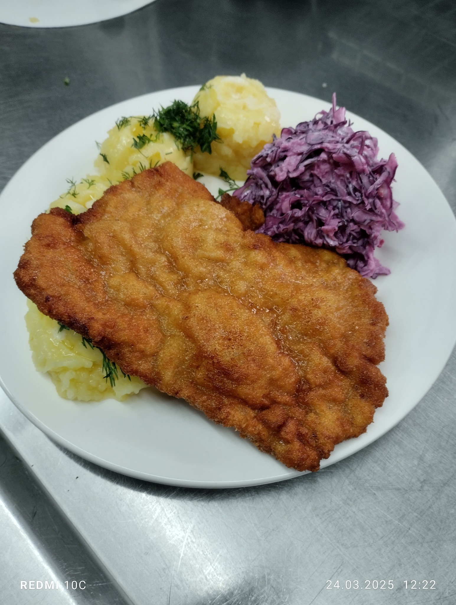 Złocisty sznycel na białym talerzu z ziemniakami i surówką z czerwonej kapusty, udekorowany koperkiem. Zdjęcie zrobione z bliska, widoczna struktura panierki.
