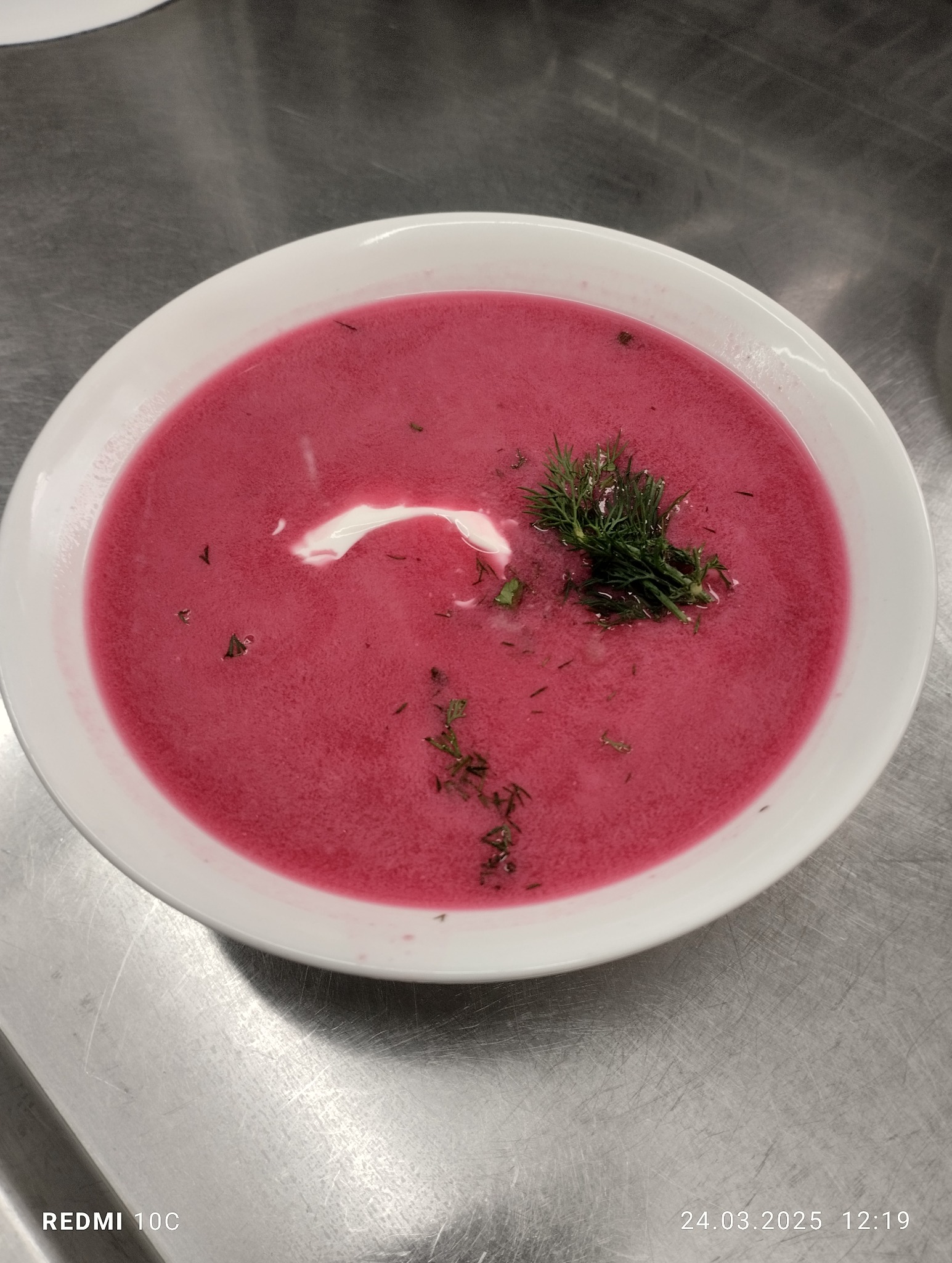 Talerz kremowego, różowego barszczu z kleksem śmietany i gałązką koperku na metalowym stole. Ujęcie z góry, naturalne światło.