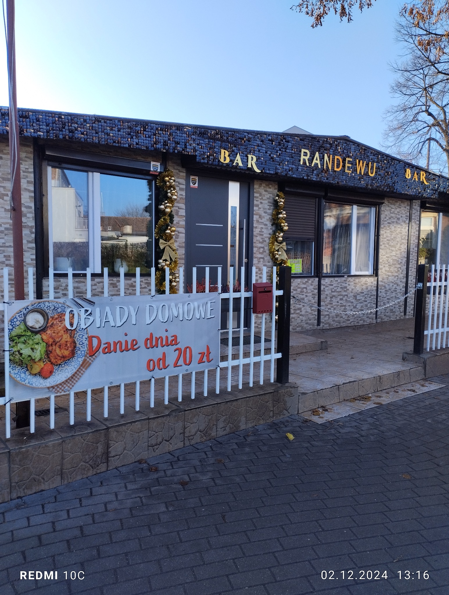 Bar Randewu z dekoracjami świątecznymi. Na ogrodzeniu reklama 'Obiady Domowe, danie dnia od 20 zł'. Widok z zewnątrz, kostka brukowa na pierwszym planie.