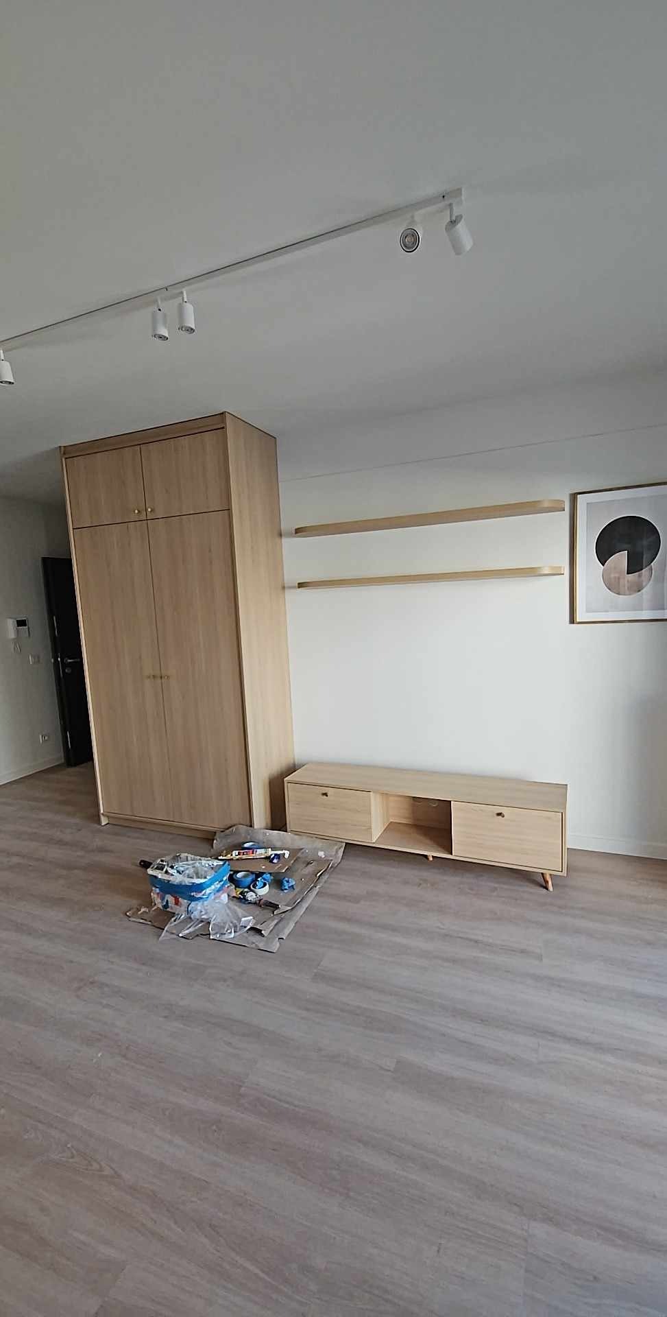 Jasne drewniane meble w pokoju: szafa, szafka RTV i półki. Na podłodze narzędzia, sufit oświetlony reflektorami. Minimalistyczny design wnętrza.