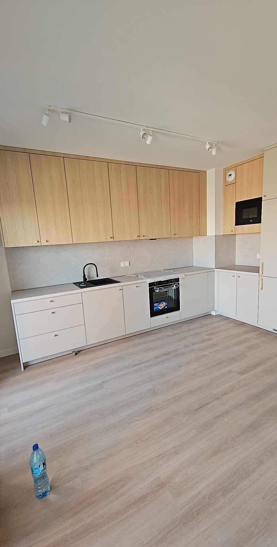 Jasna kuchnia z drewnianymi szafkami górnymi i białymi dolnymi. Na podłodze panele. Widoczna butelka wody. Oświetlenie punktowe.