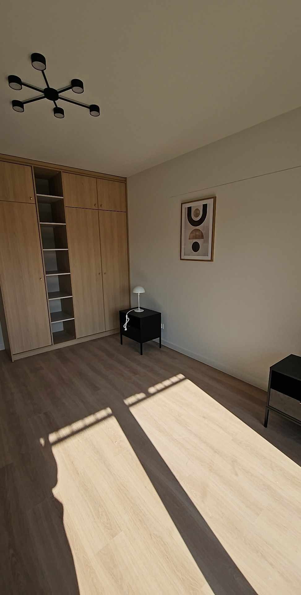 Jasne panele podłogowe w pokoju z dużą szafą w zabudowie, czarnym stolikiem nocnym i lampką. Na ścianie wisi obrazek w ramce. Widoczne cienie.