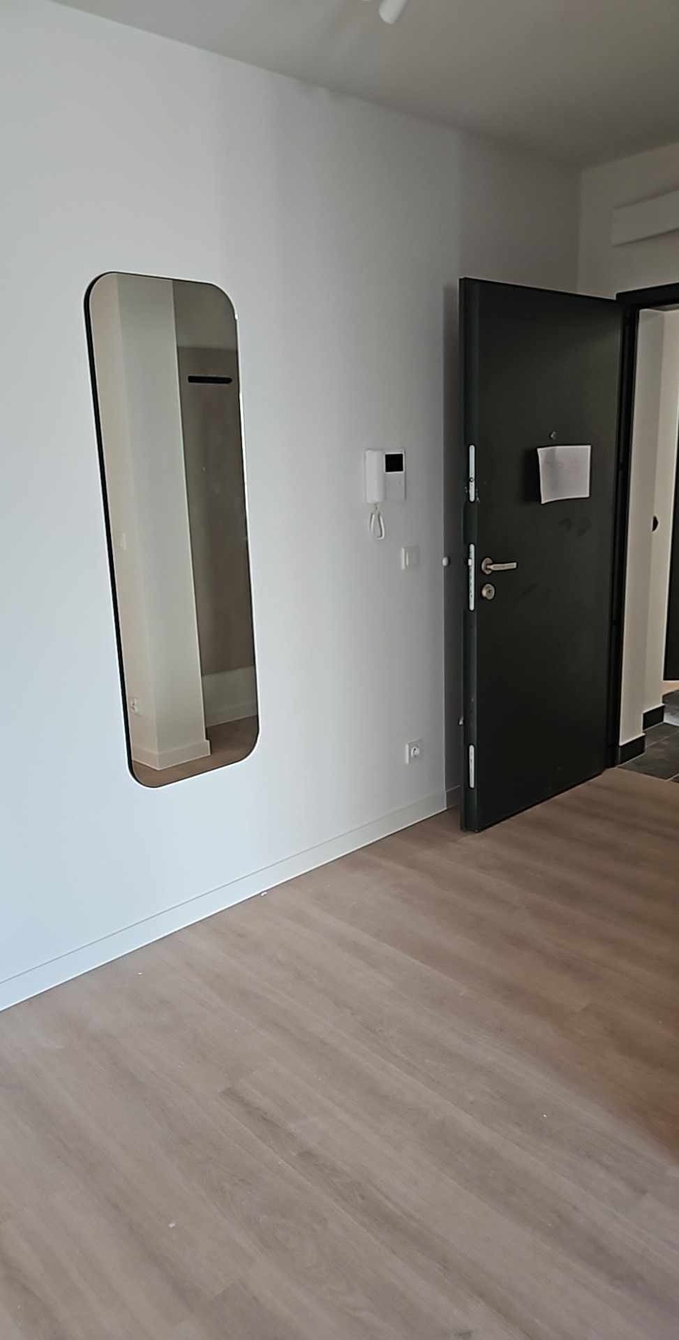 Wnętrze po remoncie: jasne panele podłogowe, białe ściany z podłużnym lustrem i czarne drzwi wejściowe z wizjerem. Minimalistyczny design.