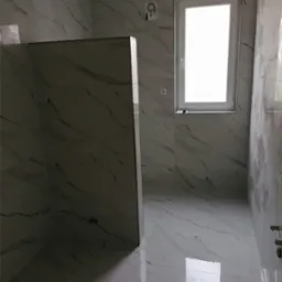 Nowoczesna łazienka z białymi płytkami imitującymi marmur, kabina prysznicowa typu walk-in z widocznymi elementami montażowymi, okno z białymi ramami, wentylator kanałowy na ścianie.