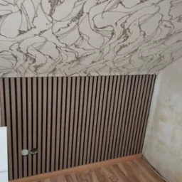 Panel ścienny z drewnianymi lamelami w brązowo-czarnych odcieniach, połączony z sufitem pokrytym wzorzystą tapetą w odcieniach beżu i brązu, widoczne gniazdka elektryczne i fragment podłogi z paneli.