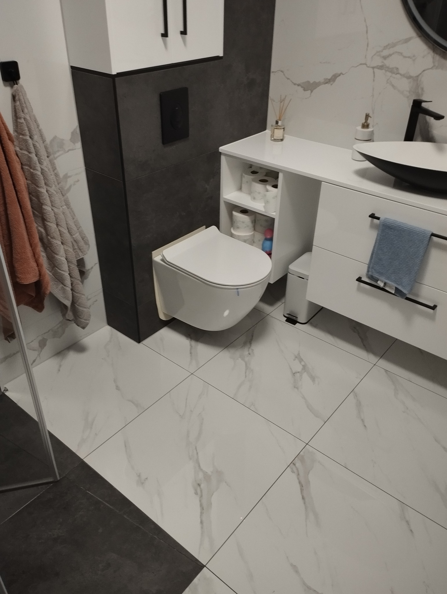 Nowoczesna łazienka z podwieszaną toaletą, białą umywalką nablatową na szafce z szufladami i marmurowymi płytkami na ścianach i podłodze.