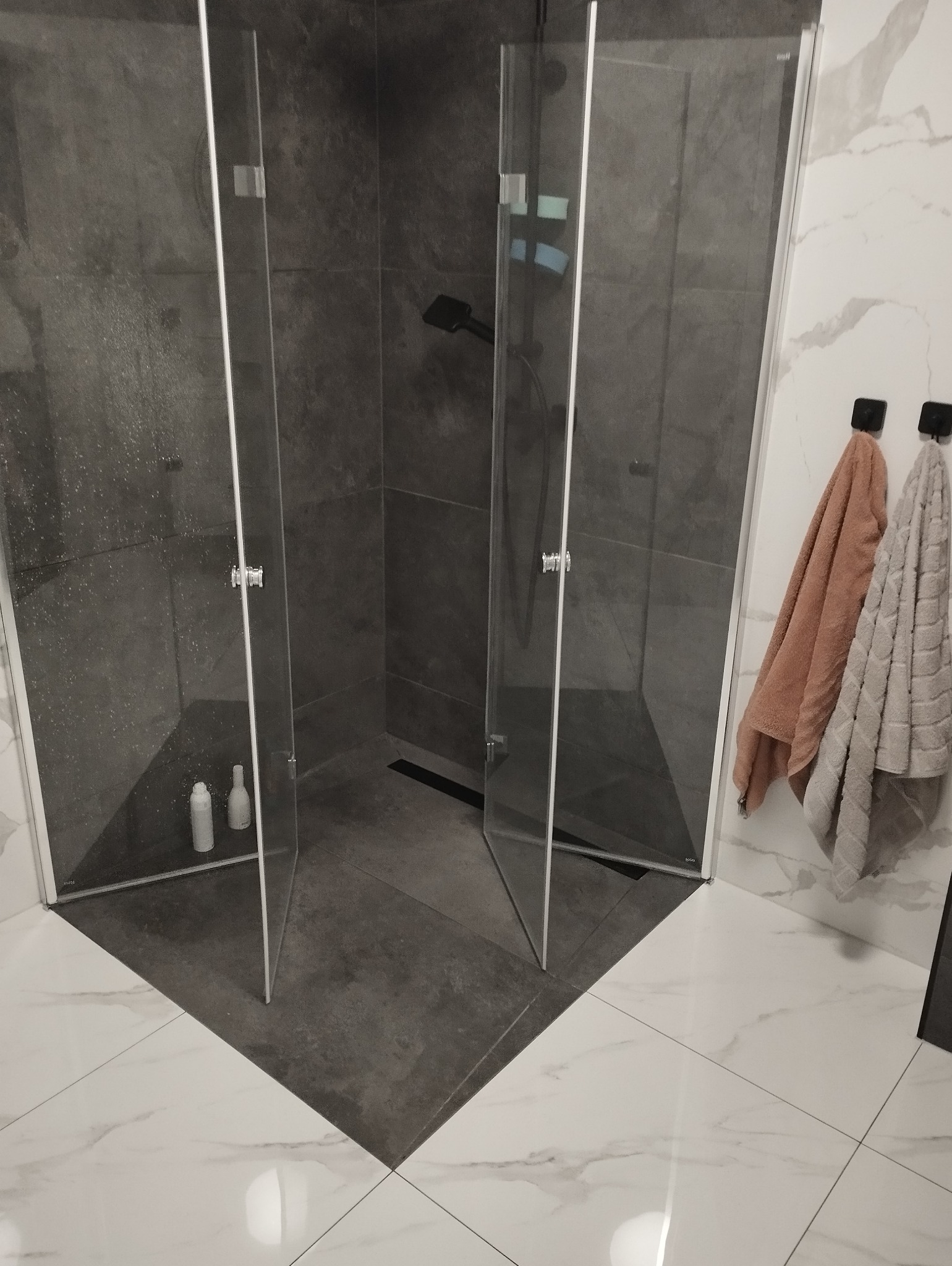Nowoczesna kabina prysznicowa z otwartymi drzwiami, szare płytki wewnątrz, białe płytki marmurowe na podłodze i ścianie, widoczne dwa ręczniki na haczykach.