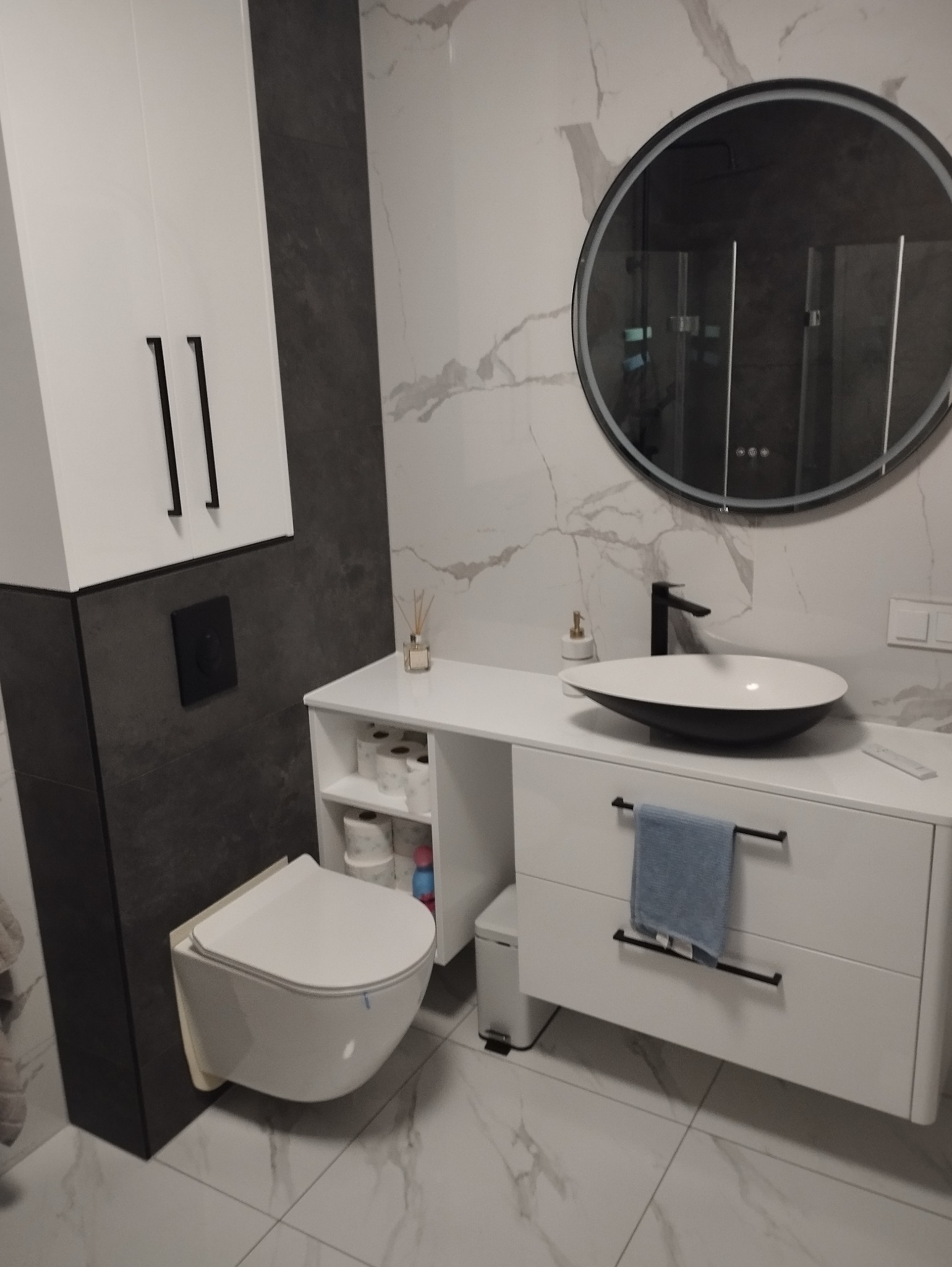 Nowoczesna łazienka z umywalką nablatową, okrągłym lustrem z podświetleniem LED, wiszącą toaletą i marmurowymi płytkami.