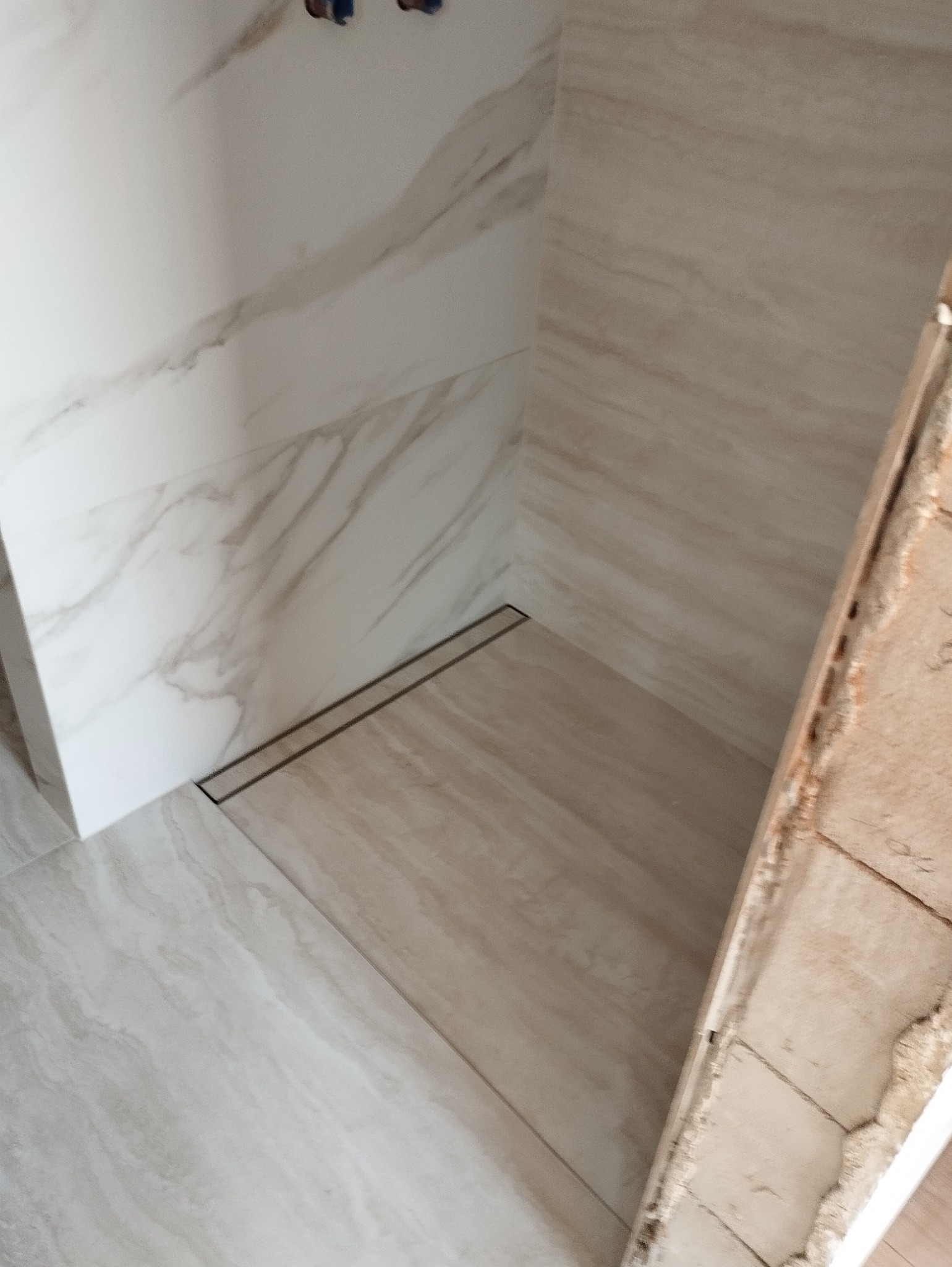 Wykończona strefa prysznicowa z odpływem liniowym i płytkami imitującymi marmur, widoczna częściowo obudowa z surowym betonem.