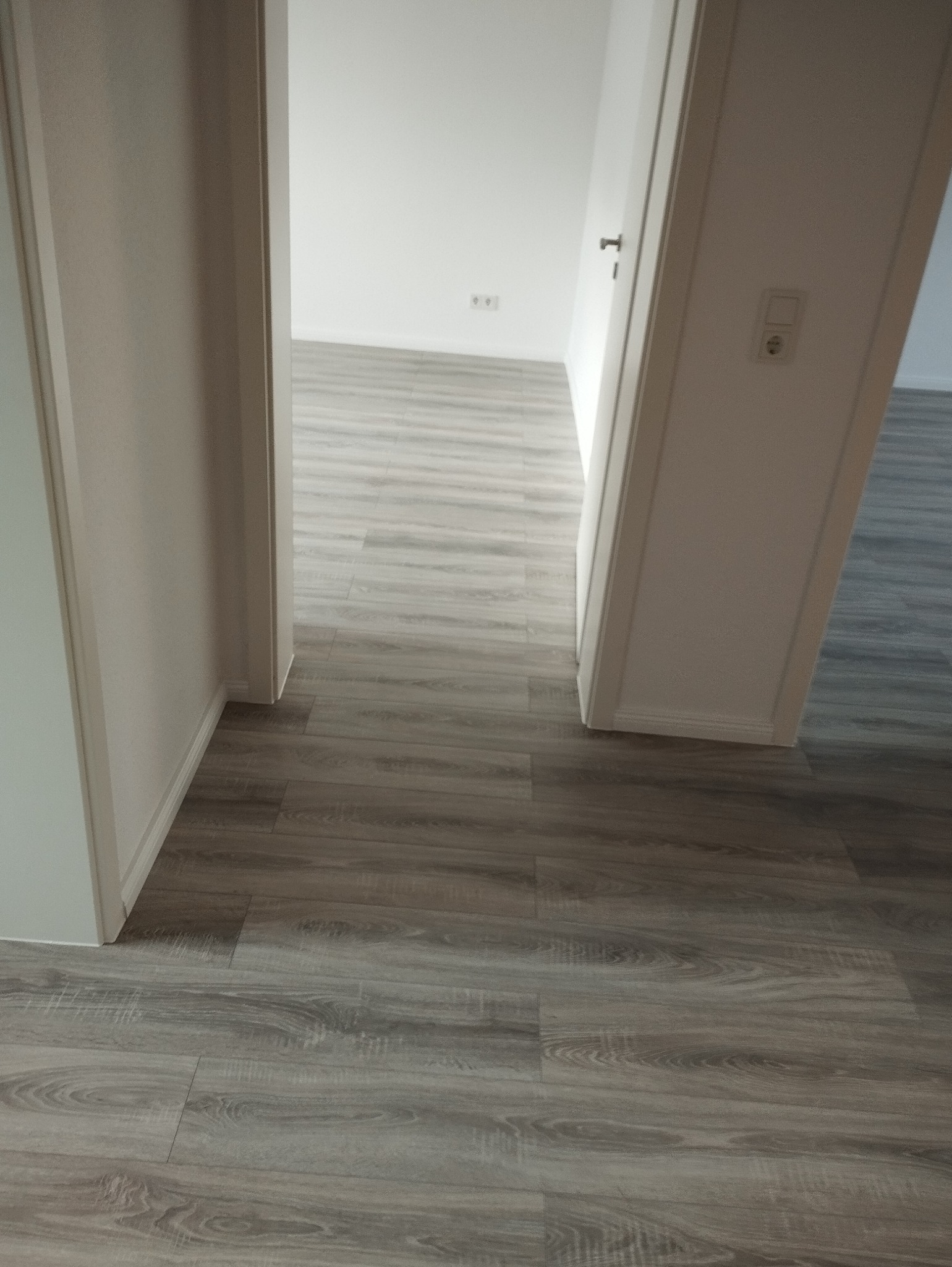 Ułożone panele podłogowe w odcieniach szarości, widoczne z perspektywy przejścia między pokojami, z białymi ścianami i listwami przypodłogowymi.