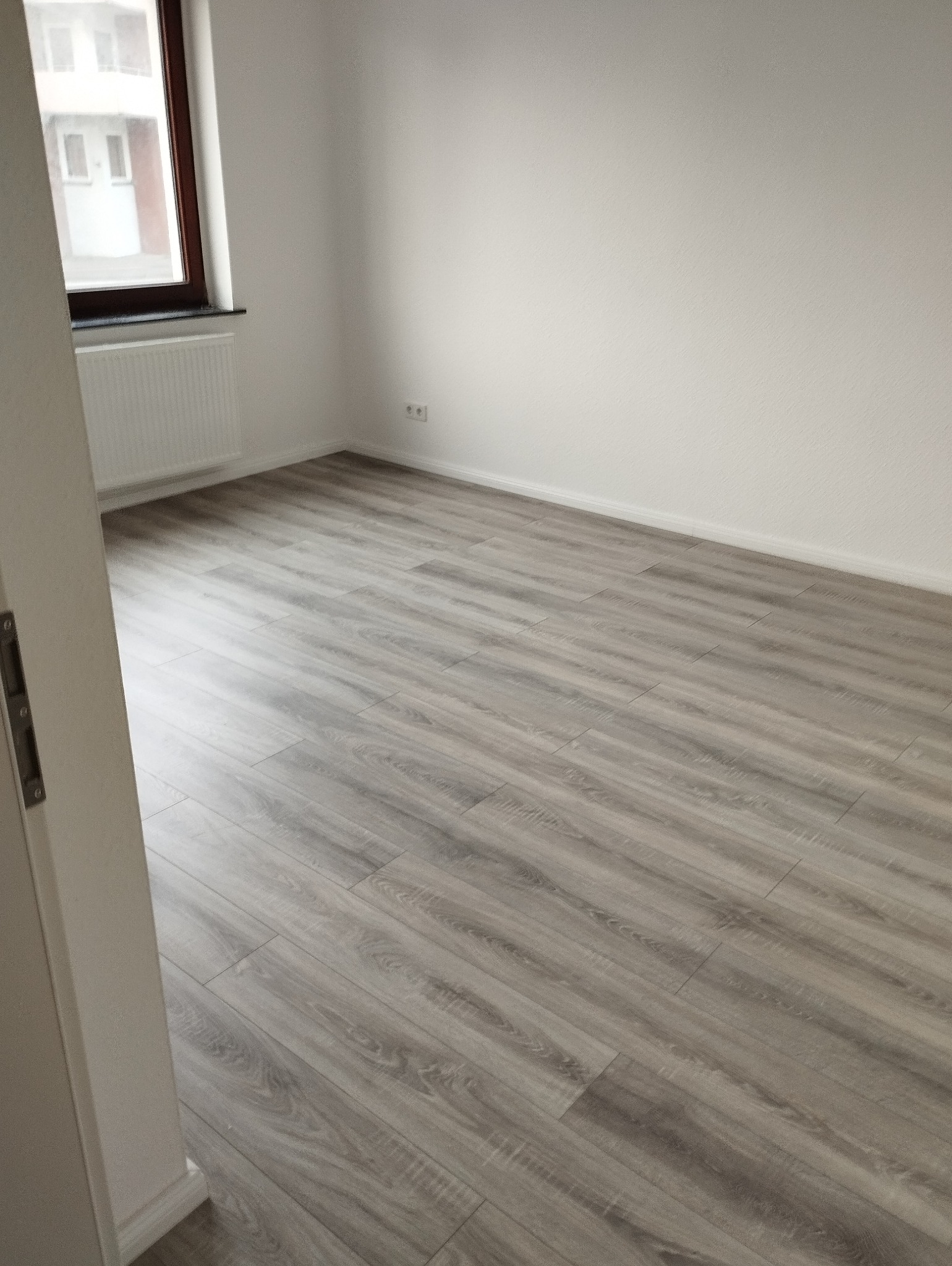 Jasnoszara podłoga z paneli ułożona w pustym pokoju z białymi ścianami, widok zza otwartych drzwi.